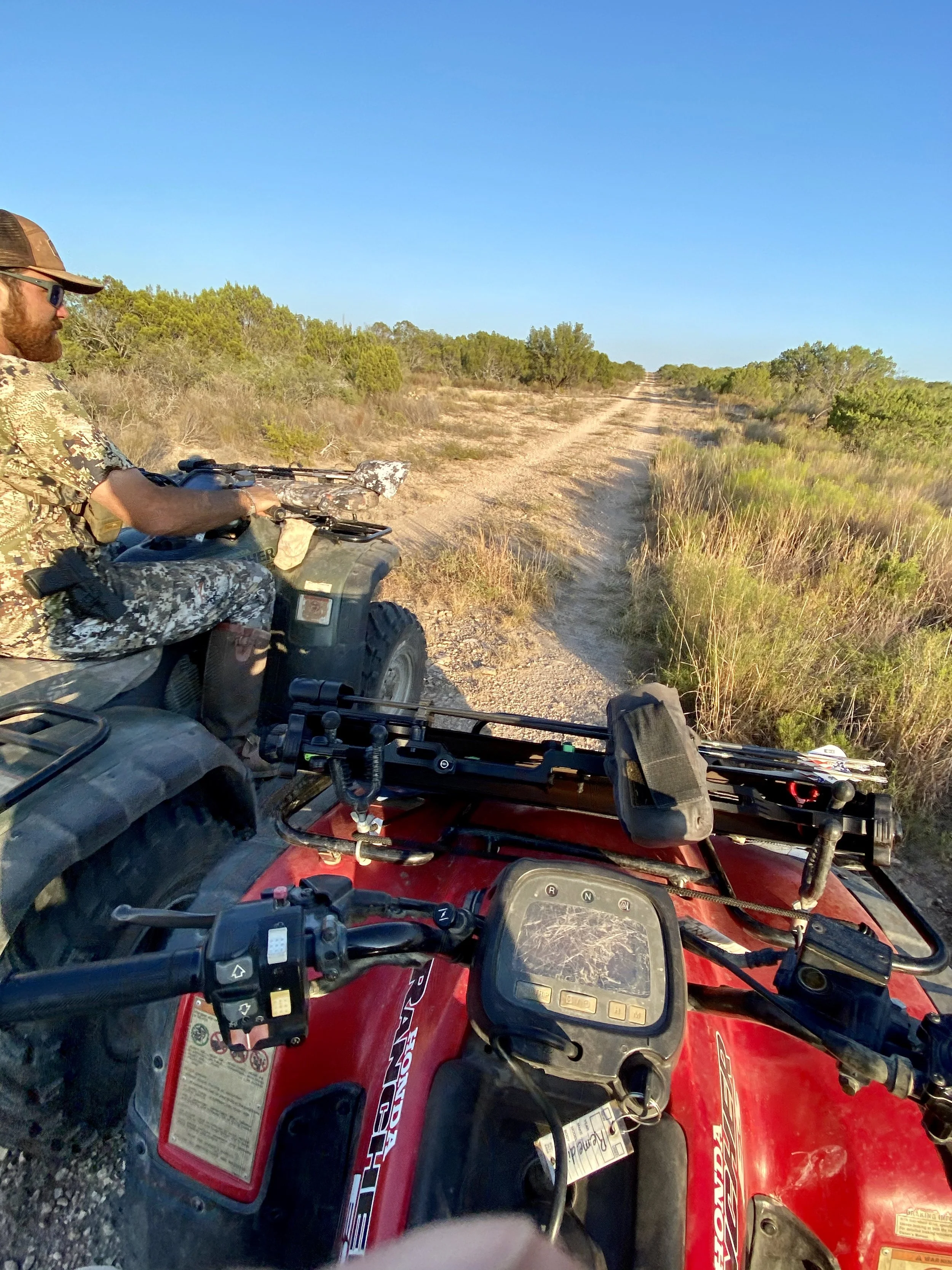 View from the windshield of a red all-terrain vehicle (ATV) on a dirt trail through a rural area with sparse, low vegetation and small trees under a clear blue sky.