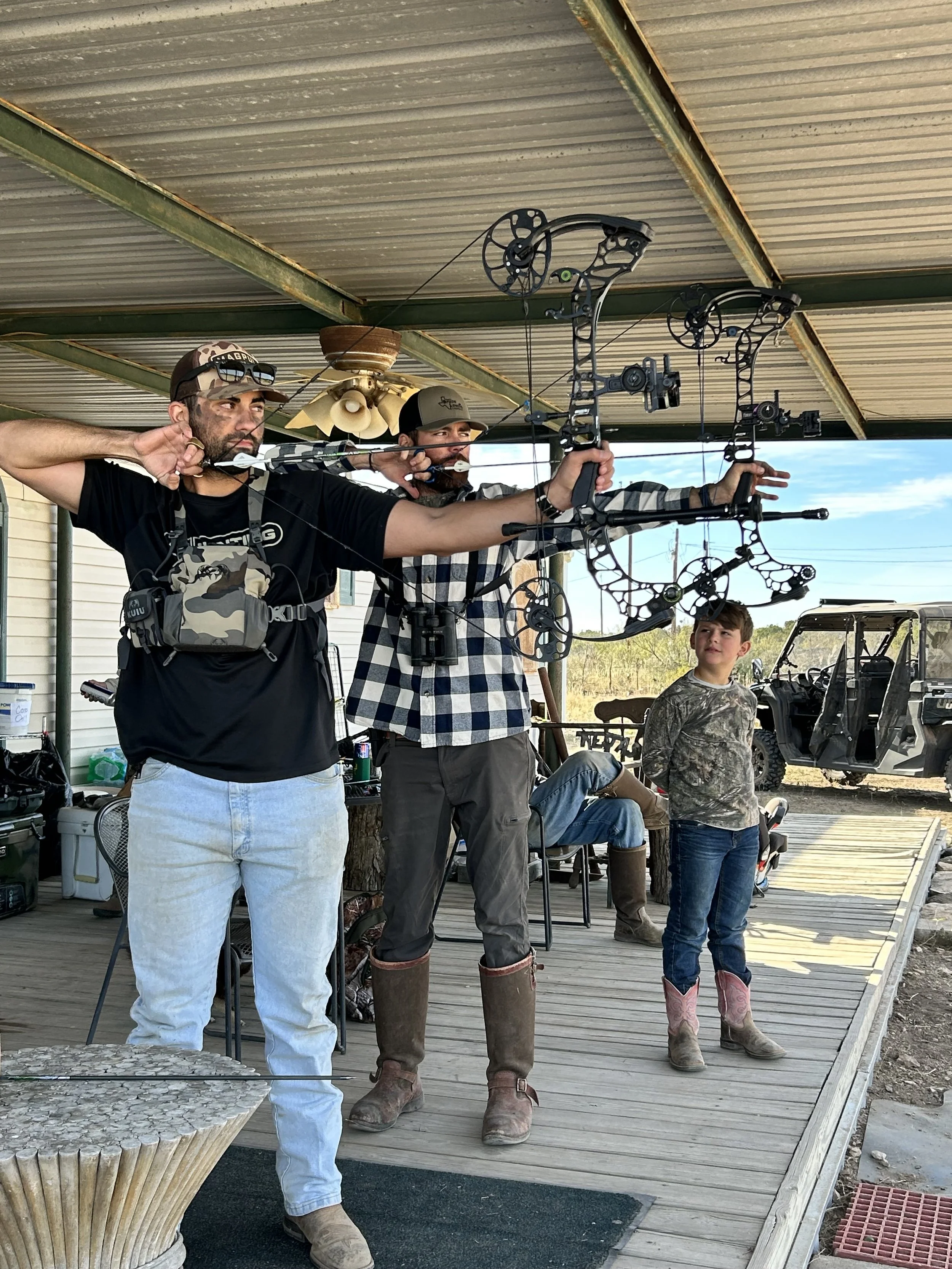 Three people aiming bows and arrows outdoors on a wooden porch. The person in the foreground is wearing a camouflage cap, sunglasses, and a black t-shirt, while the person in the middle is wearing a gray cap, camouflage shirt, and tall boots. A young
