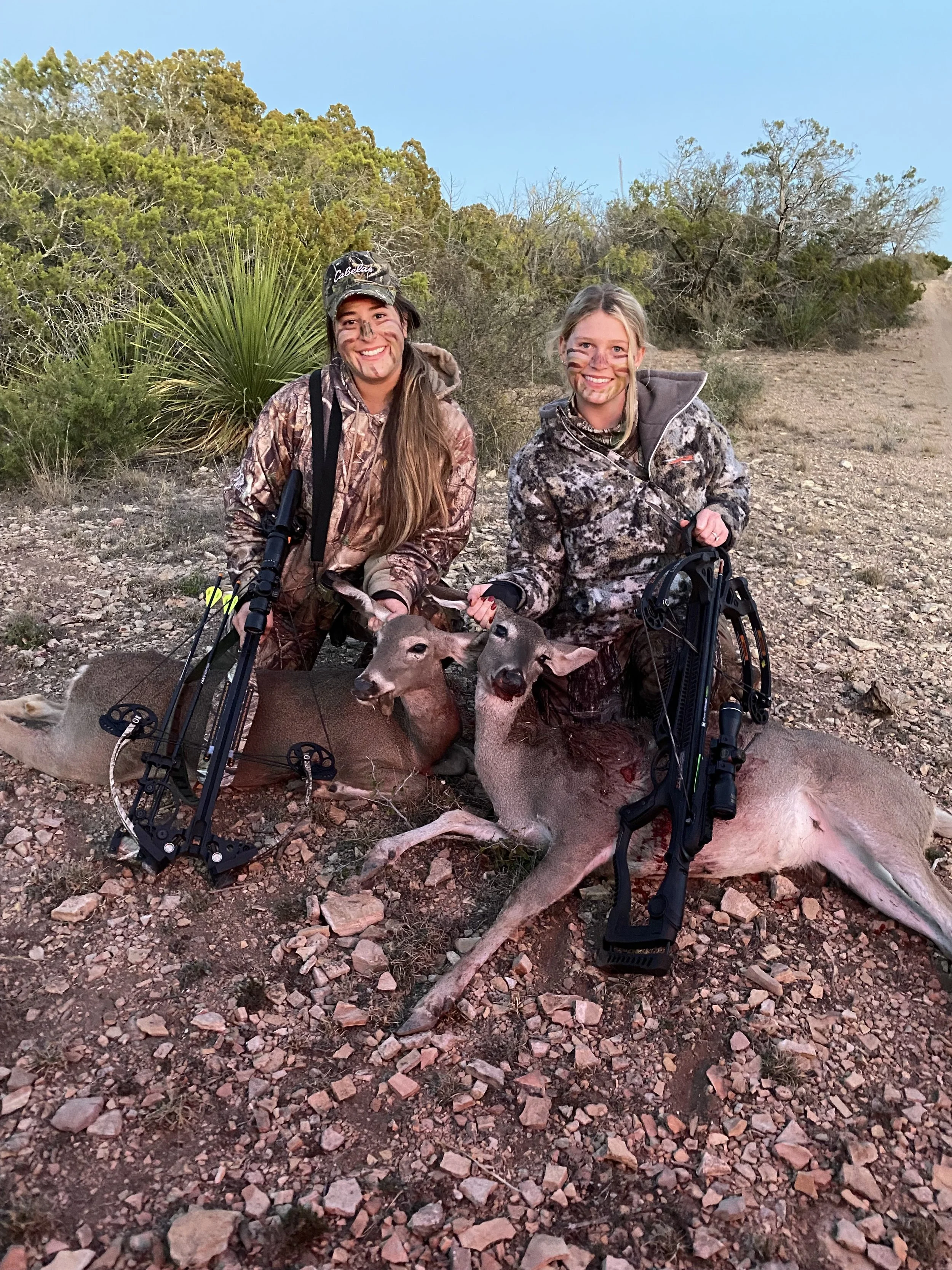 Two women in camouflage hunting clothes kneel on rocky ground, smiling, with two deer they appear to have hunted, and both hold bows. The background has desert shrubbery and a clear blue sky.