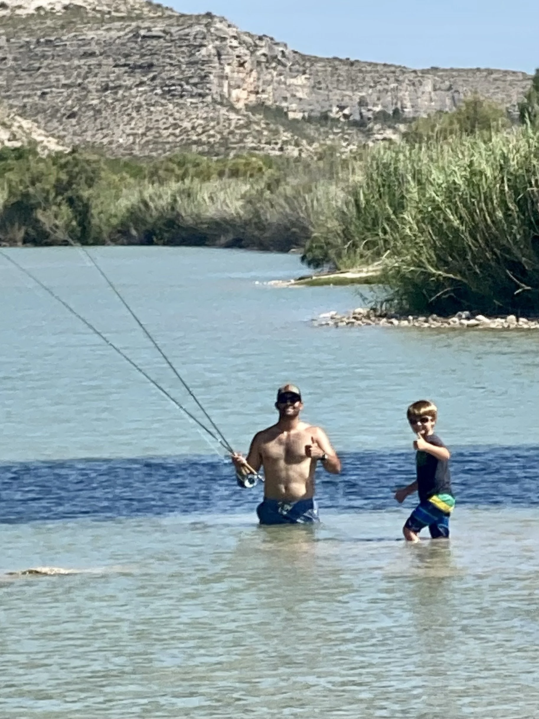 Two males, one adult and one young boy, standing in shallow water at a lake with a rocky hillside and sparse vegetation in the background. The adult male is holding a fishing rod and is shirtless, while the boy is wearing a black shirt and colorful s