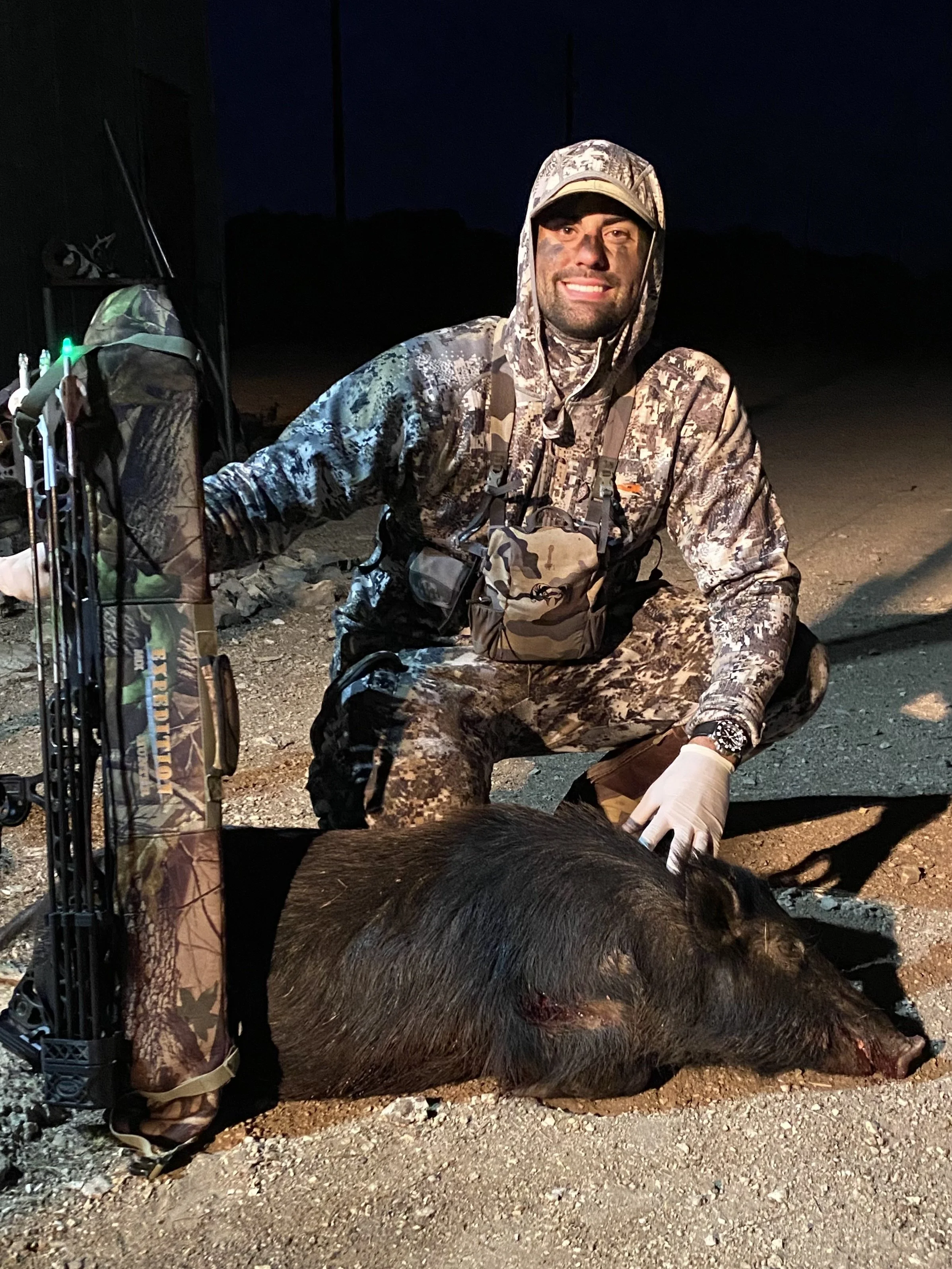 A man in camouflage hunting gear kneeling next to a large hunted pig on the ground at night.