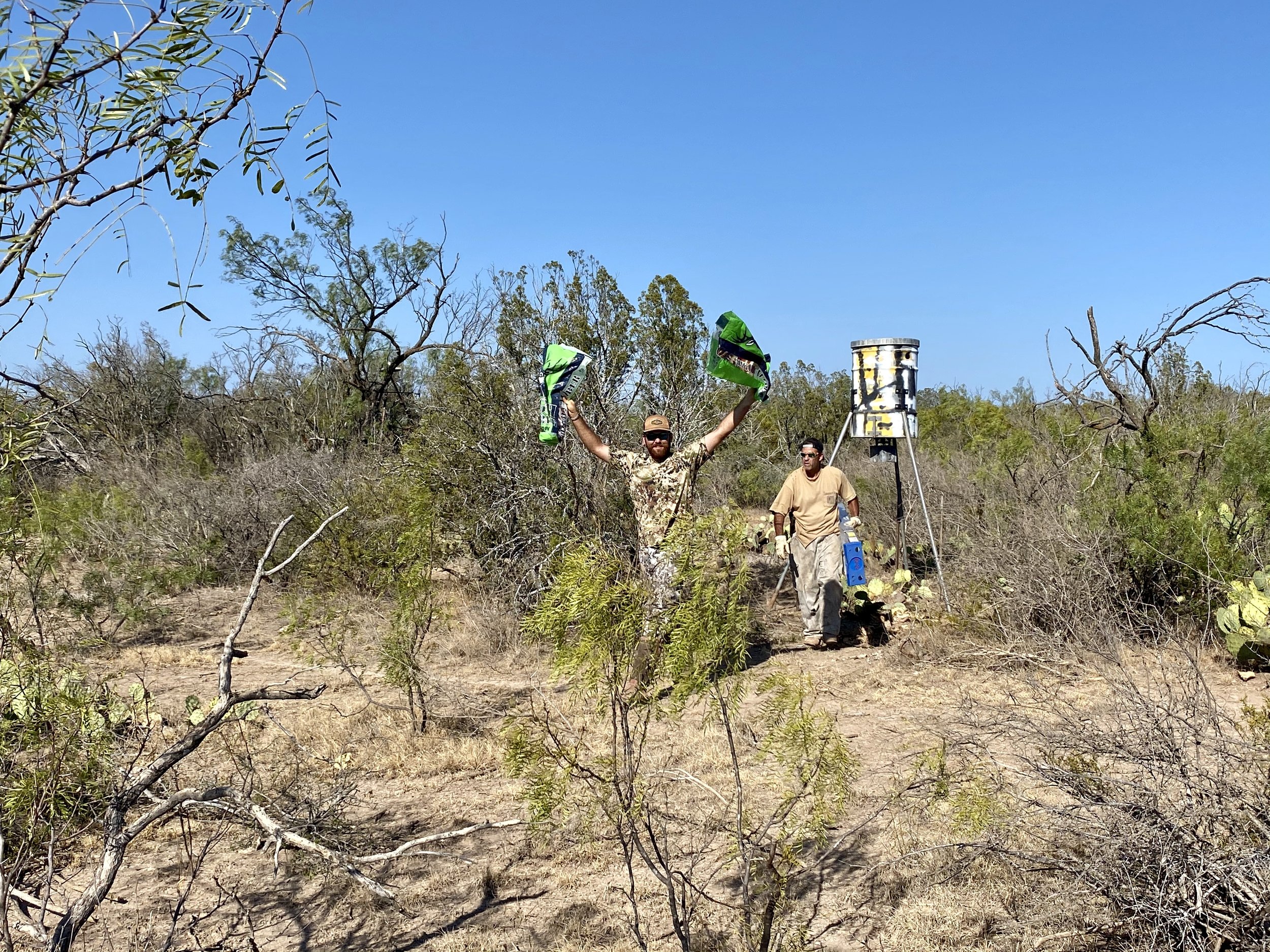 Two men in a dry, shrubby area are setting up a scientific or wildlife tracking device, with one holding flags and the other near equipment, under a clear blue sky.
