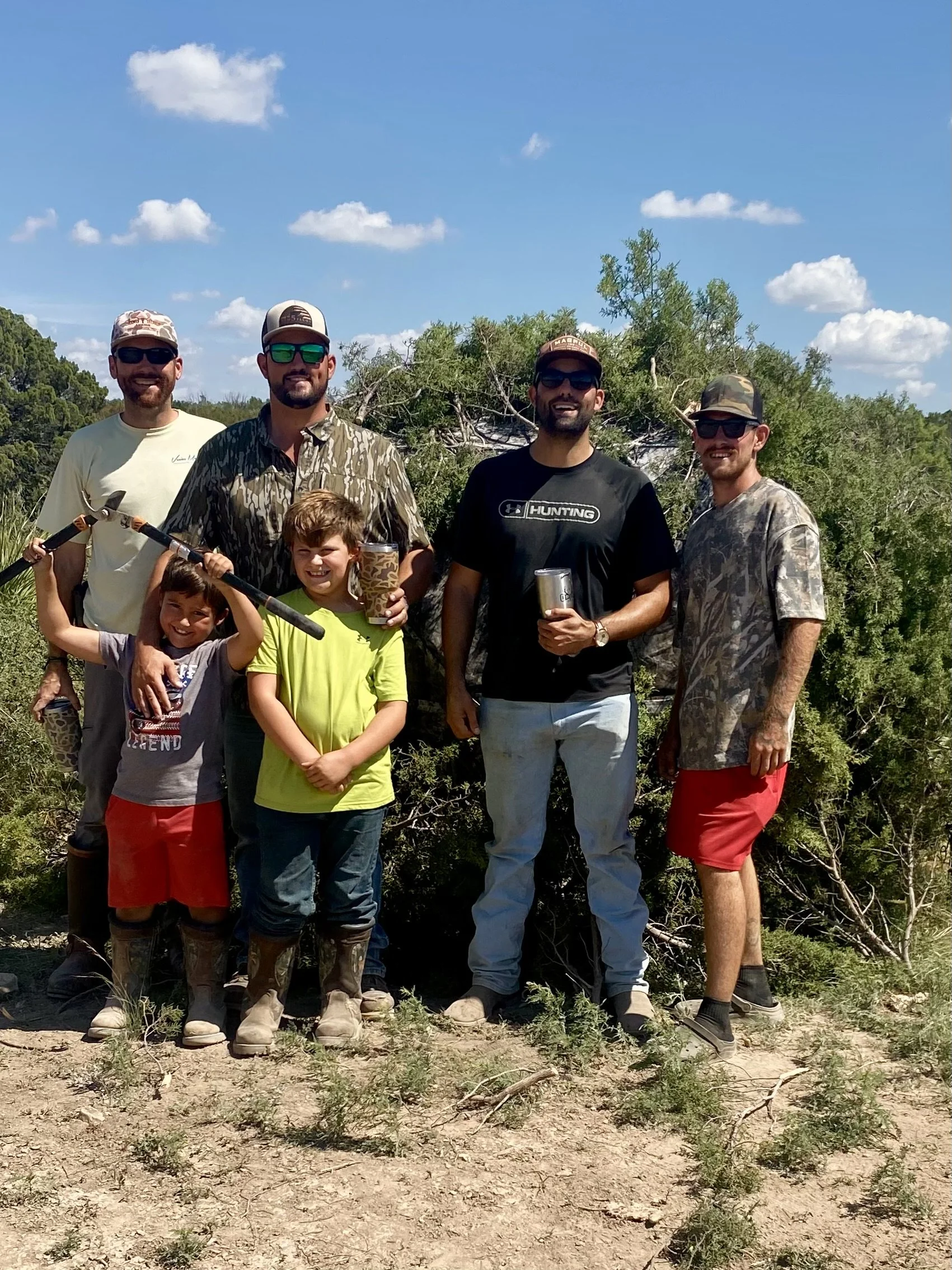 Group of six people, three adults and three children, standing outdoors in a natural setting with bushes and trees, under a blue sky with scattered clouds, smiling and holding drinks and equipment.