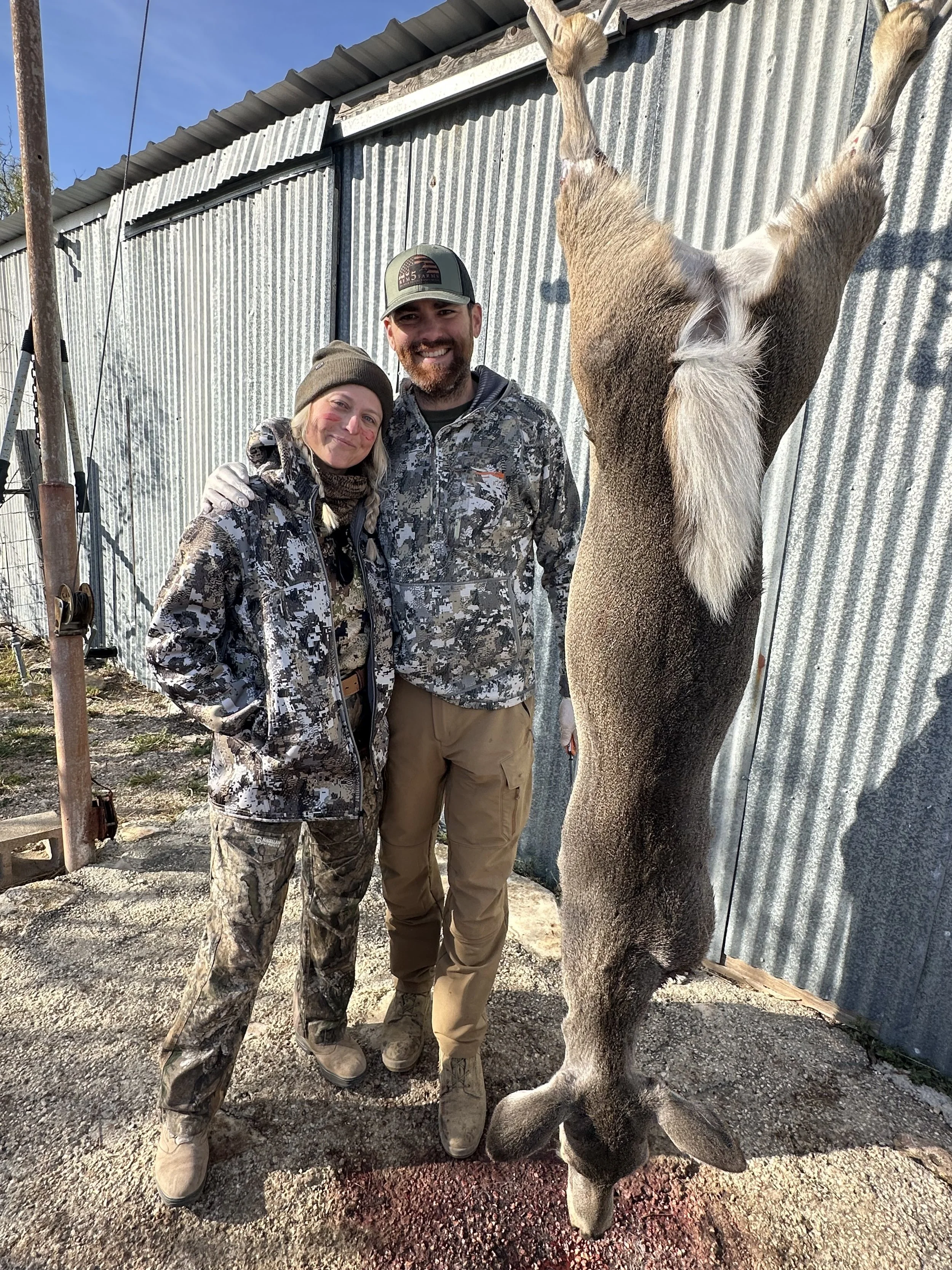 Two people dressed in camouflage outdoor clothing smiling and standing next to a kangaroo, which is facing away from the camera, outdoors near a metal building.