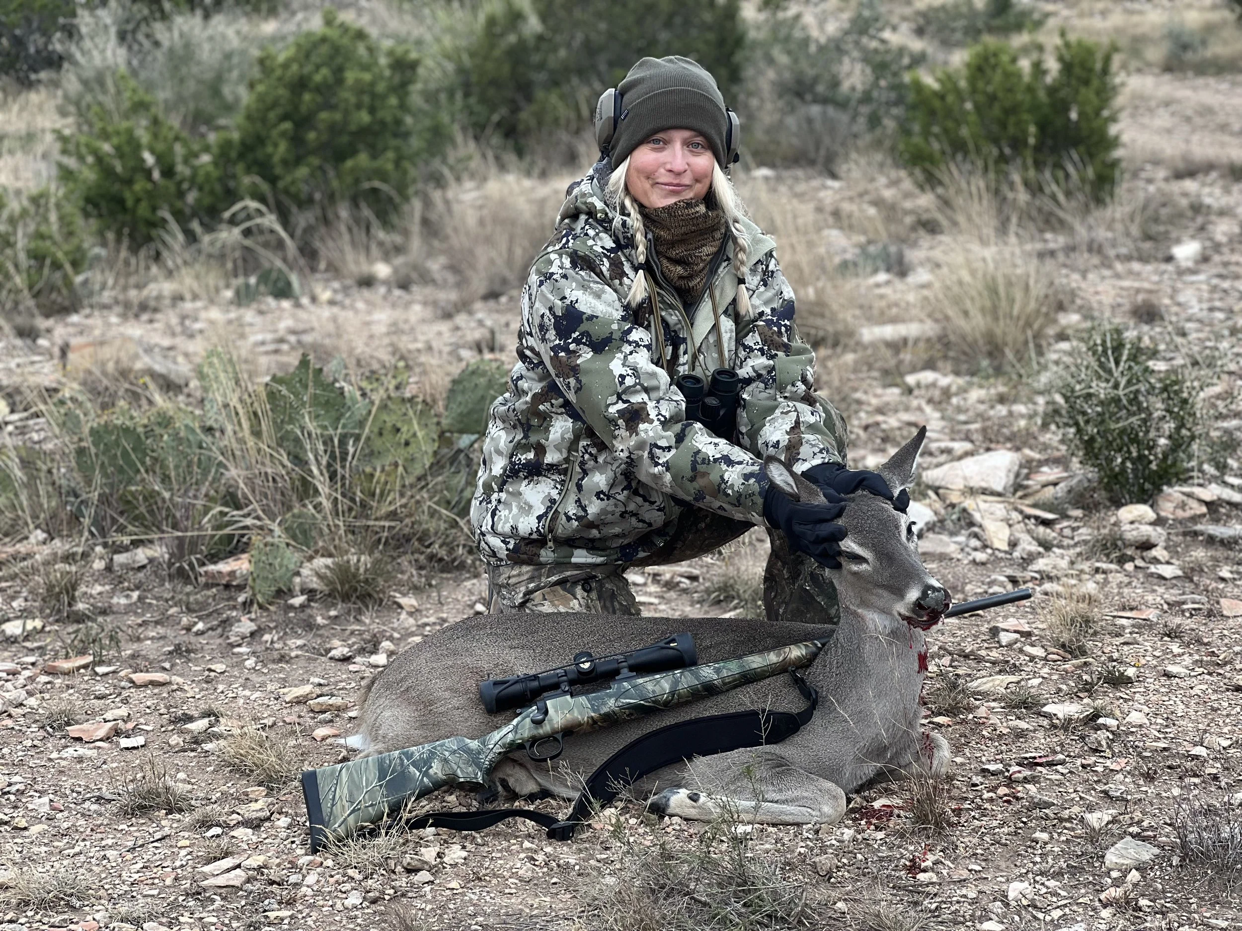 A woman dressed in camouflage hunting gear sits on the ground outdoors next to a large dead animal, holding its head. A rifle and binoculars rest on the animal, and the landscape features dry grass, rocks, and green bushes.