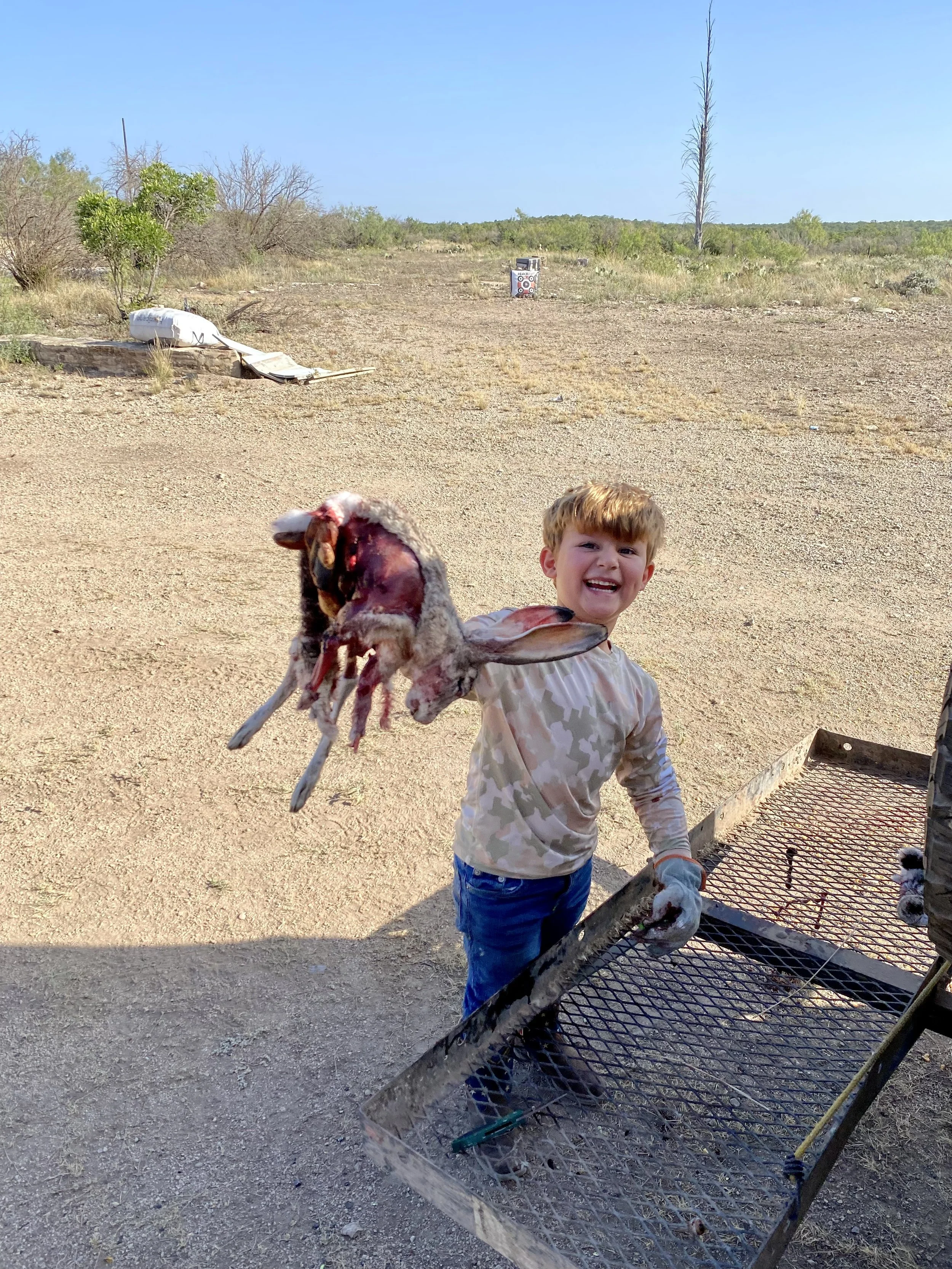 Young boy holding a freshly slaughtered sheep, standing outdoors in a dry, rural area with sparse vegetation and a target in the background.