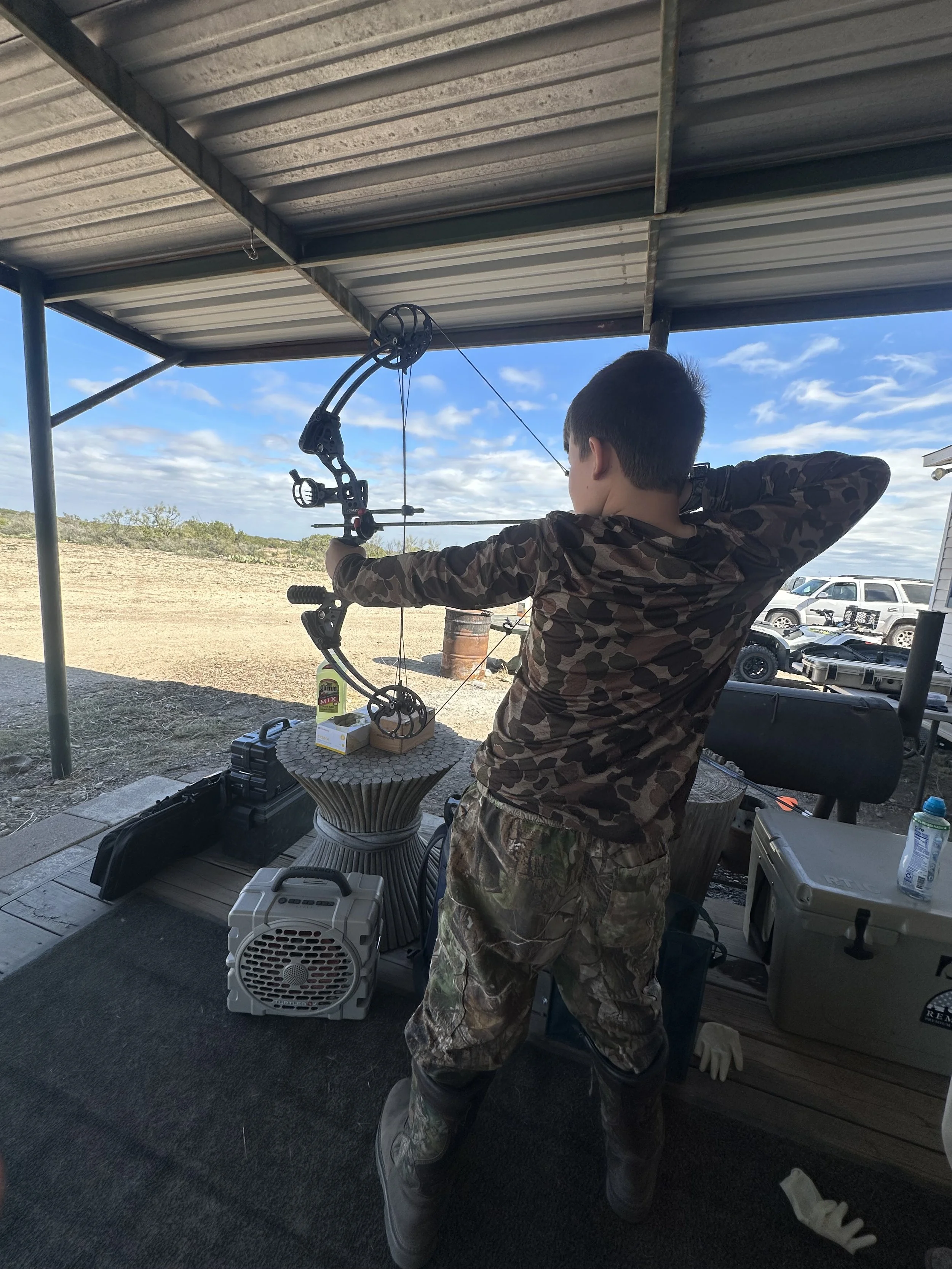 A person wearing camouflage clothing aiming a bow and arrow at an outdoor shooting range under a metal roof.