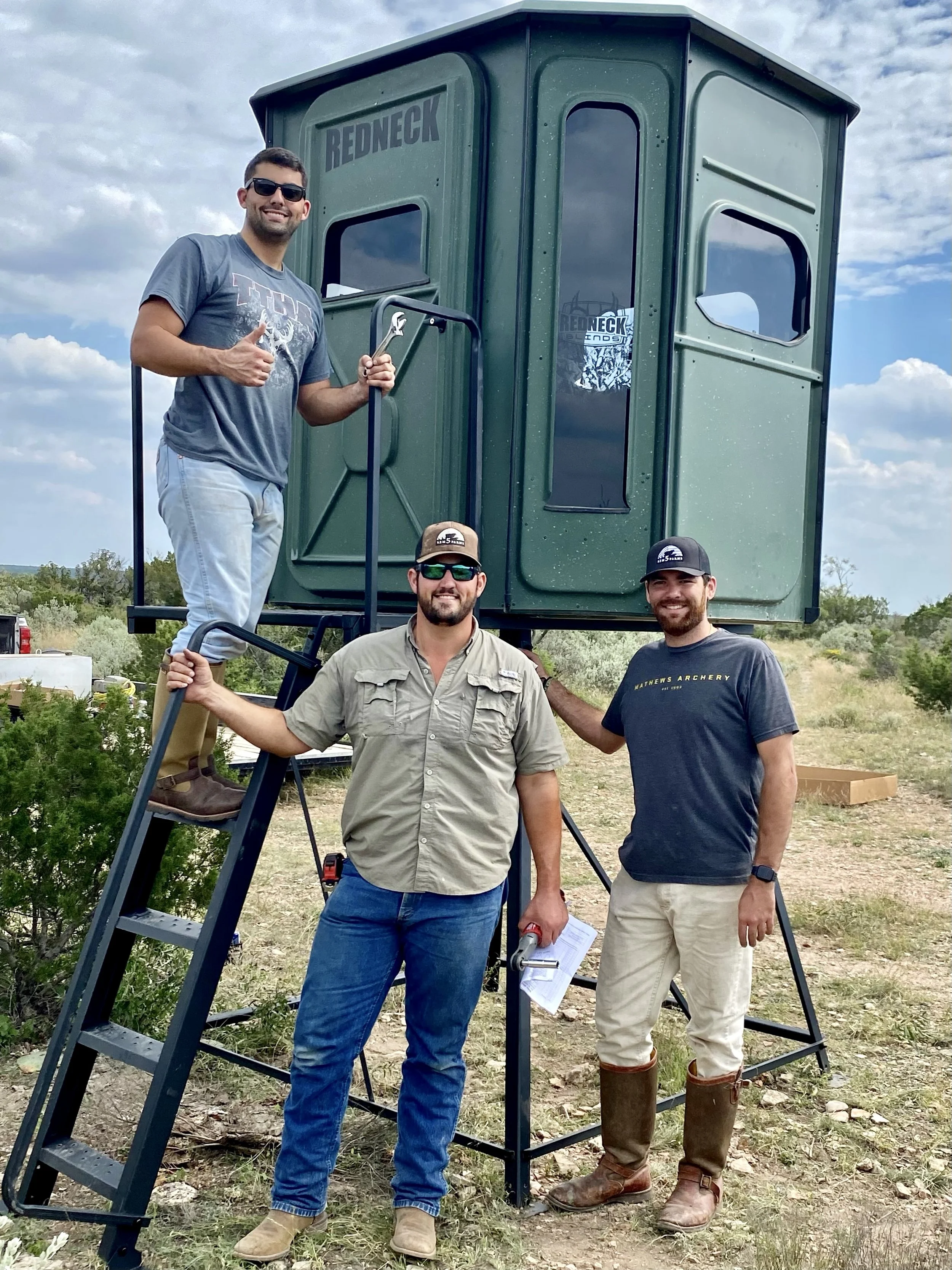 Four men standing outside in a field with a green hunting blind labeled "Redneck," some are giving thumbs up or holding papers, and one is holding a wrench.
