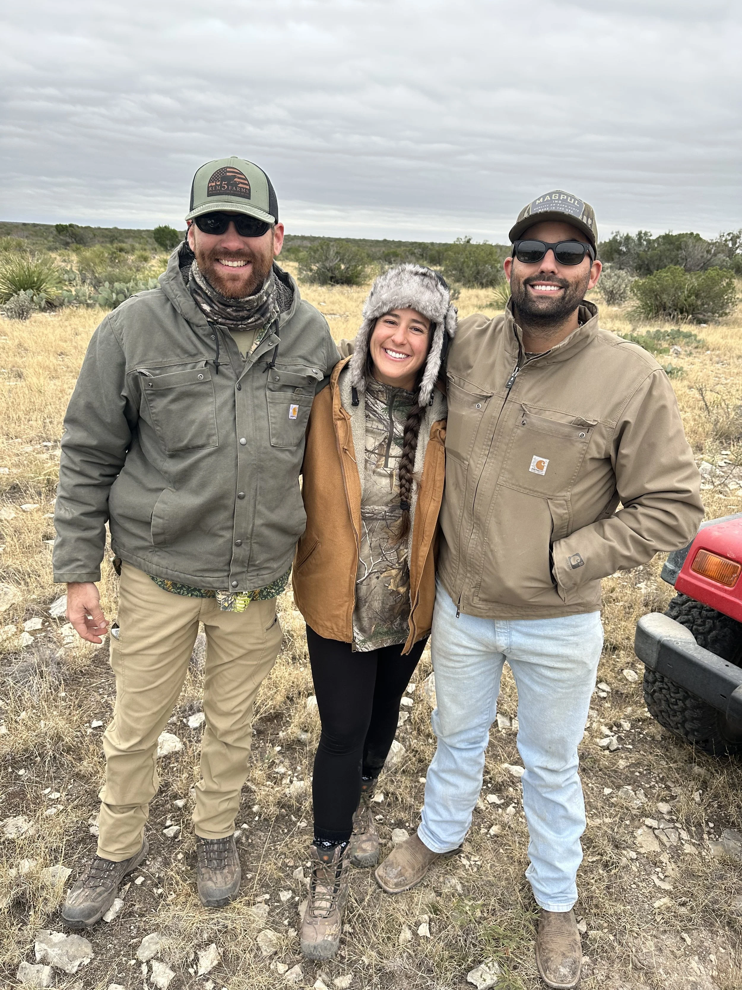 Three smiling people standing outdoors in a desert landscape with shrubs and cacti, wearing outdoor clothing and sunglasses.