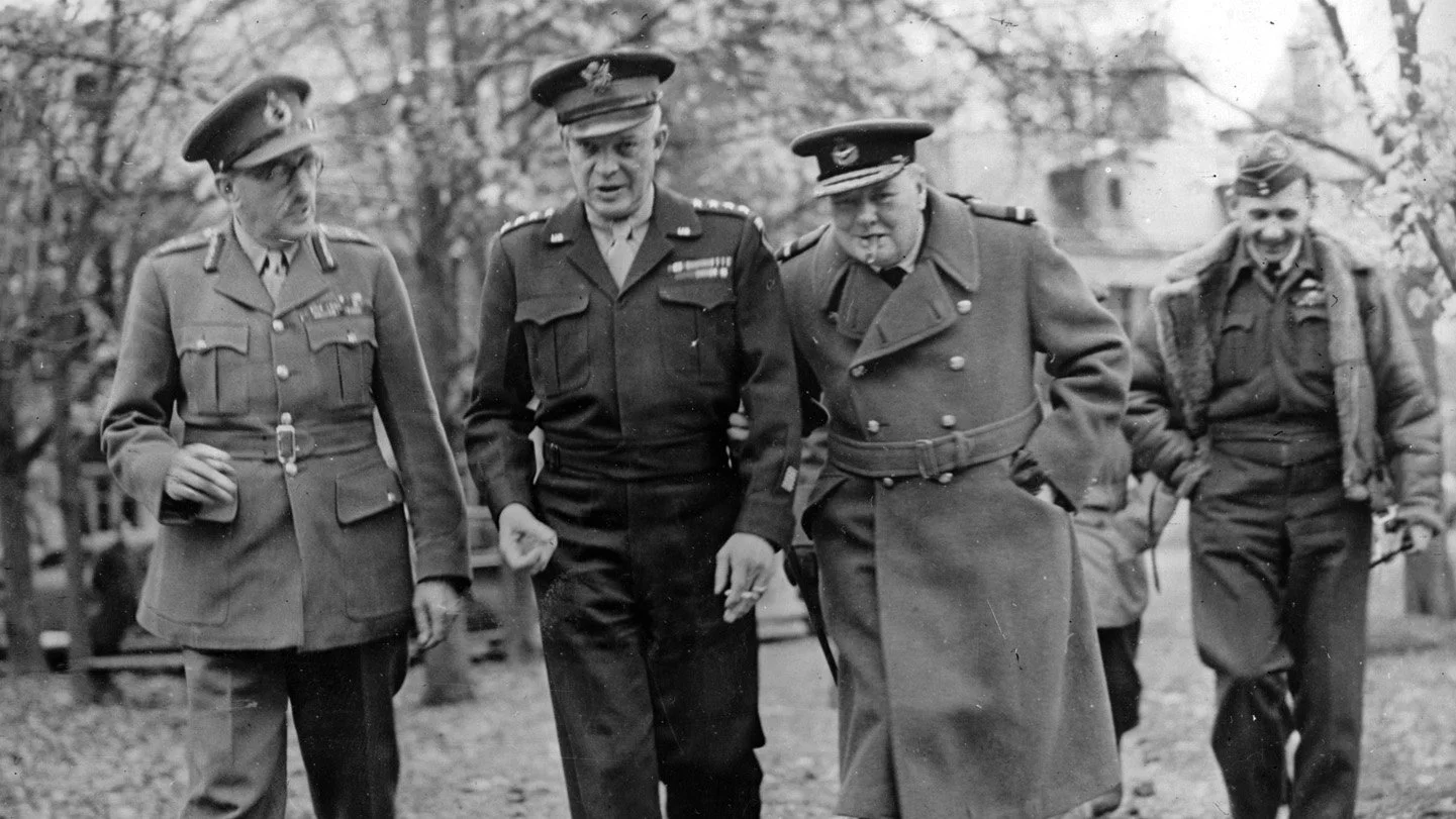 Black and white photo of four military officers in uniform walking outdoors, with trees and buildings in the background. They are engaged in conversation, smiling and gesturing.