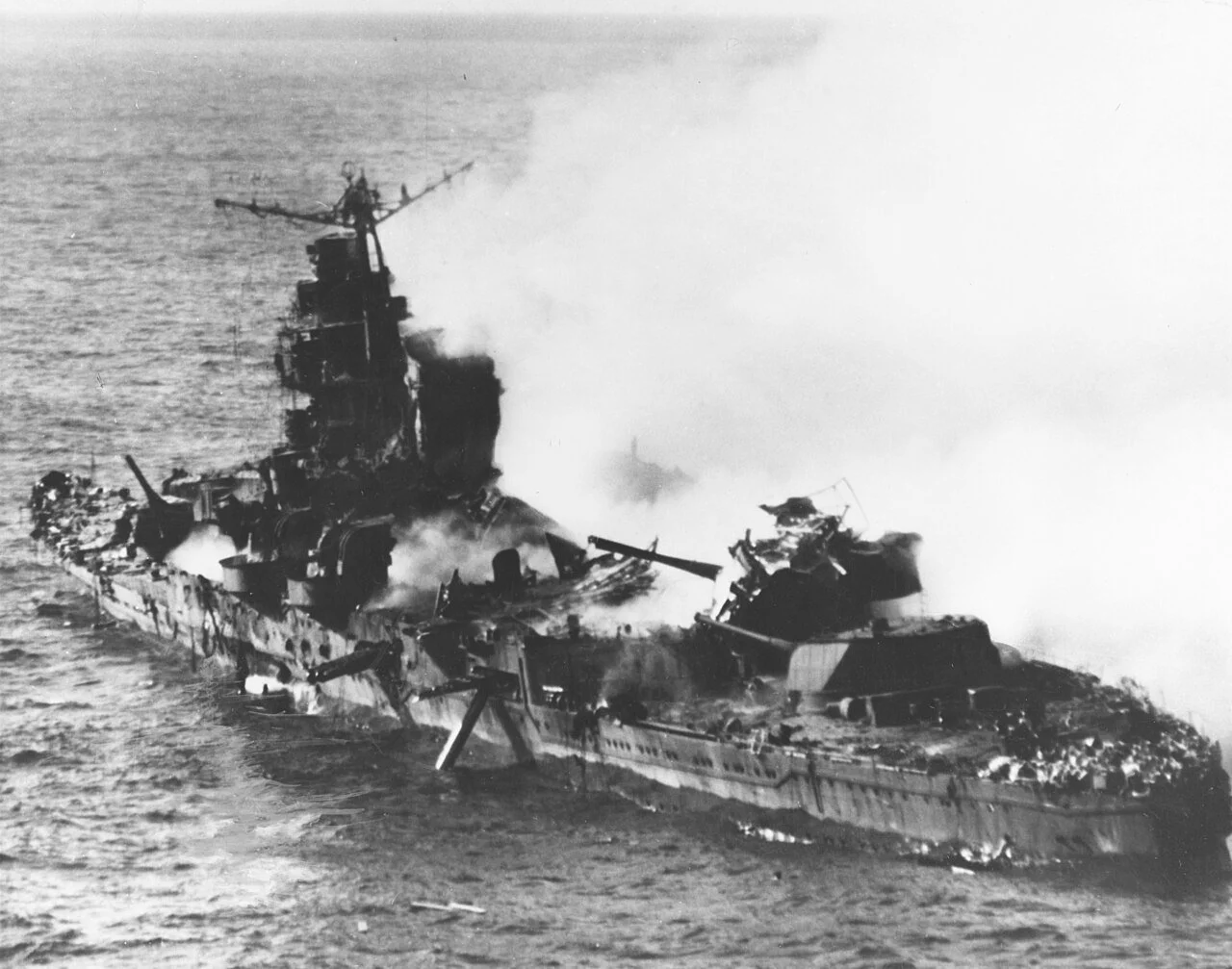 Black and white photograph of a large battleship on fire, with smoke and damage visible on the ship's deck and superstructure, in the ocean.