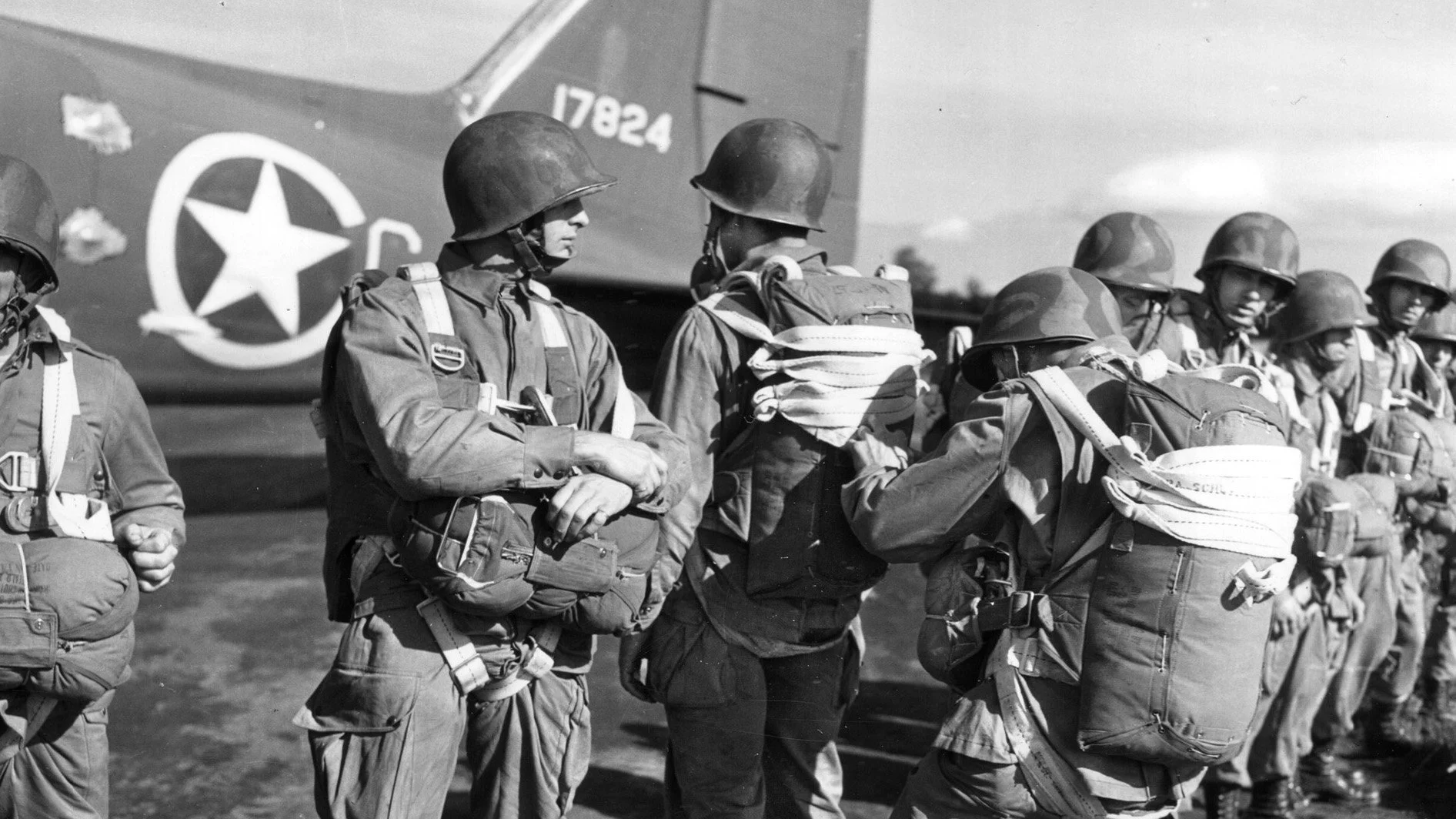 Black and white photo of soldiers in uniform and helmets standing in a line with a military aircraft in the background.