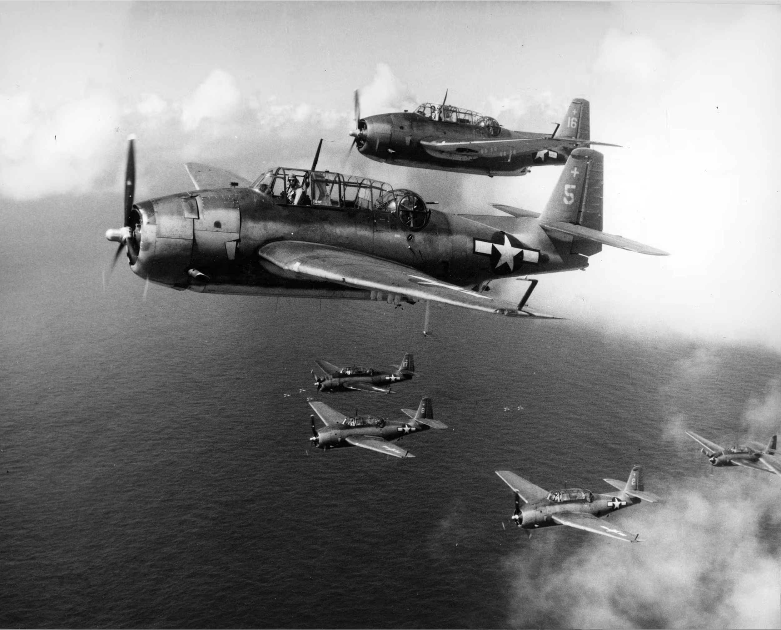 Black and white photograph of World War II aircraft flying in formation over the ocean. The aircraft have military markings and are flying at a high altitude with clouds in the background.