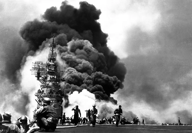 Black and white photo of soldiers and military personnel on a ship's deck with large explosions and thick black smoke billowing in the background.