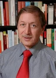 Portrait of a man in a light blue shirt and red tie, standing in front of a bookshelf filled with books.