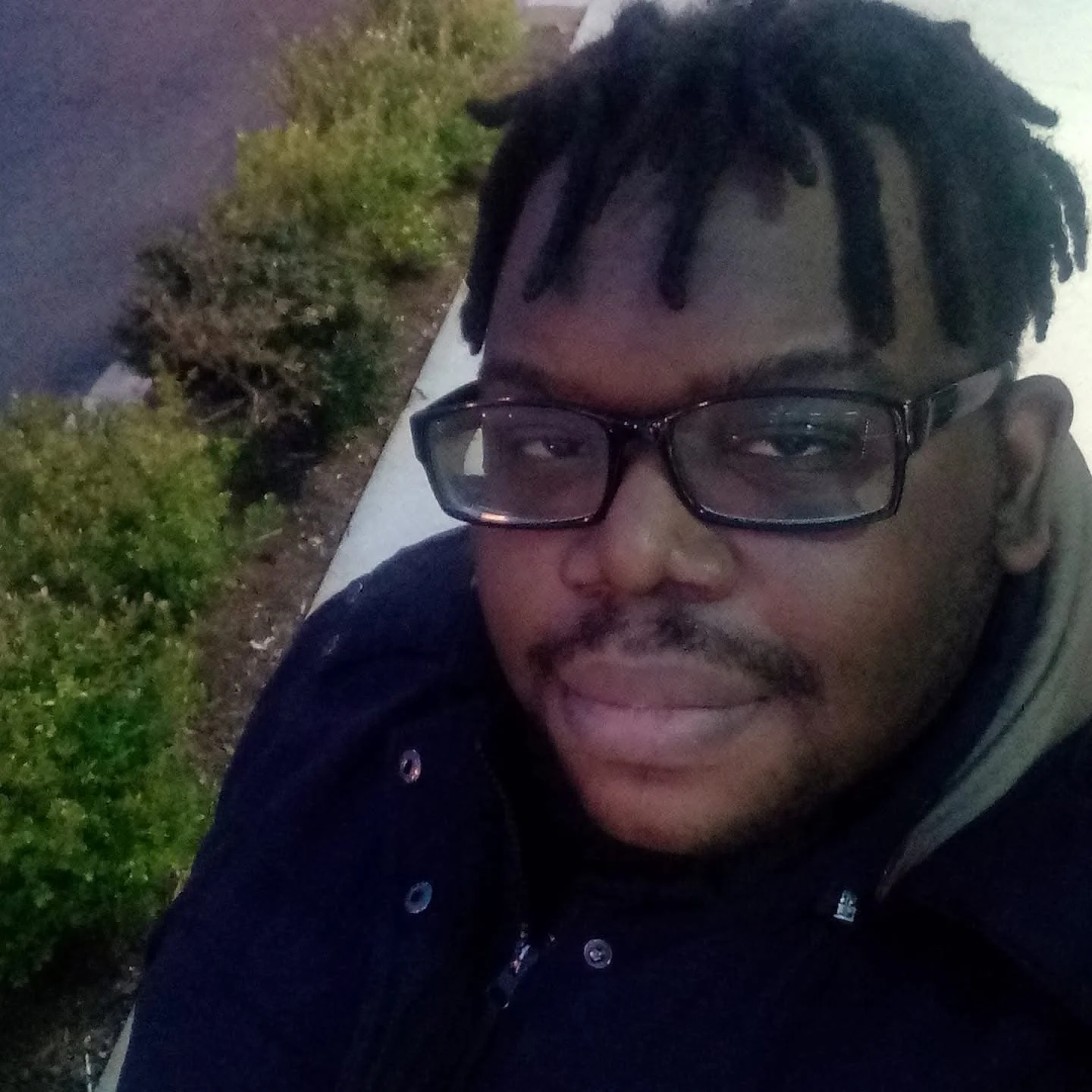 A man with glasses and dreadlocks taking a selfie outdoors in the evening, with bushes and a pathway visible in the background.