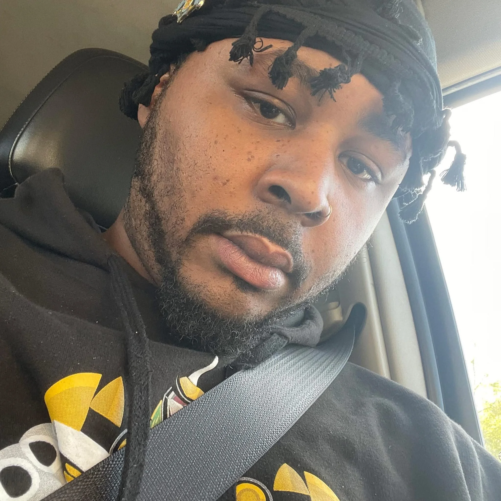 Close-up of a man with dark skin, facial hair, and dreadlocks, wearing a dark hoodie and sitting in a vehicle seat near a window.