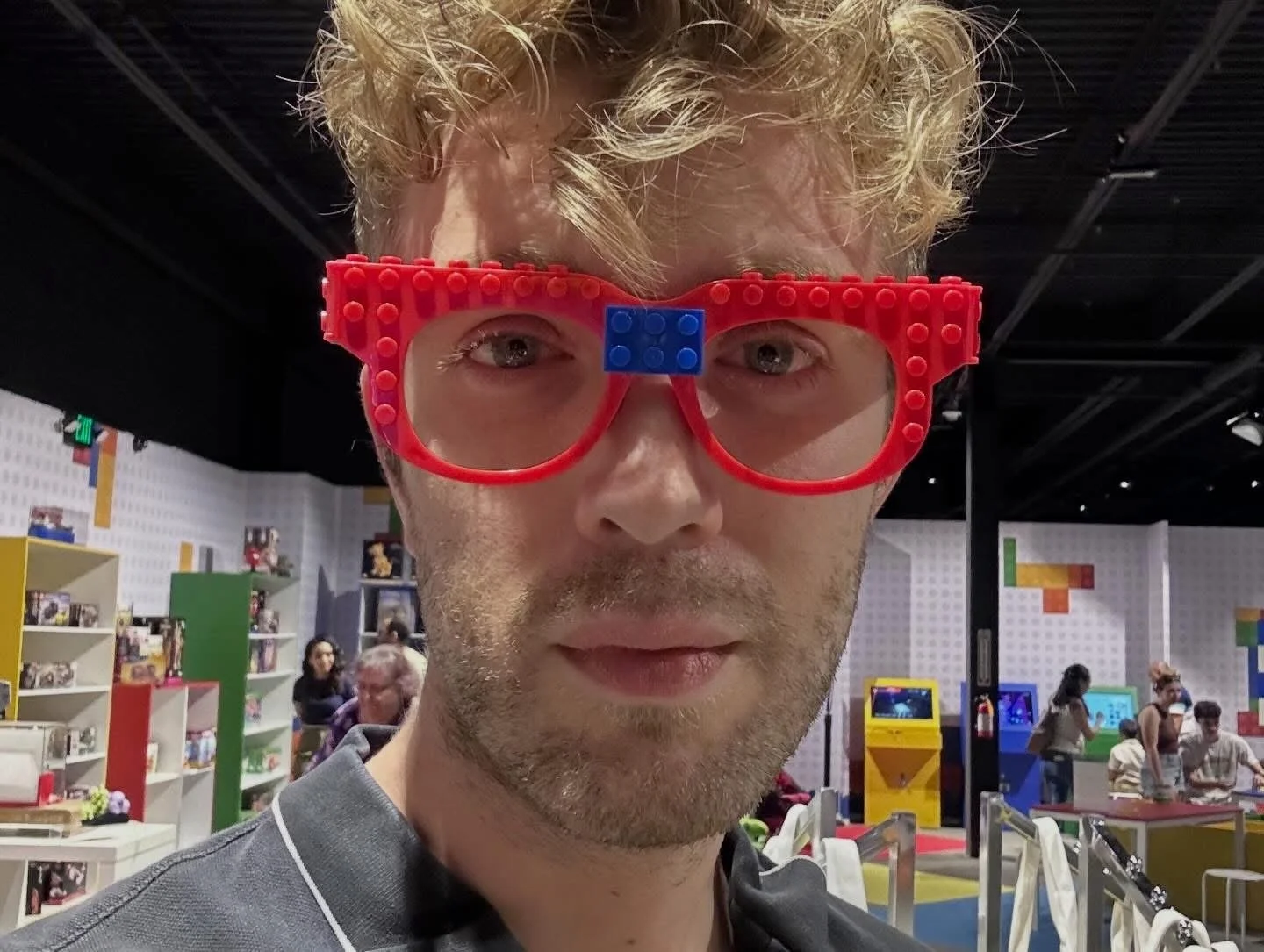 A young man with blonde curly hair wearing large red glasses with a blue brick in the middle, standing in a colorful indoor amusement or game center, with shelves and other people in the background.