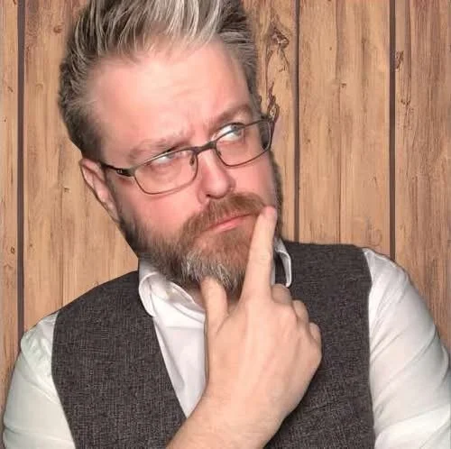 A man with glasses and a beard, wearing a gray vest over a white shirt, poses contemplatively with his hand on his chin, against a wooden panel background.