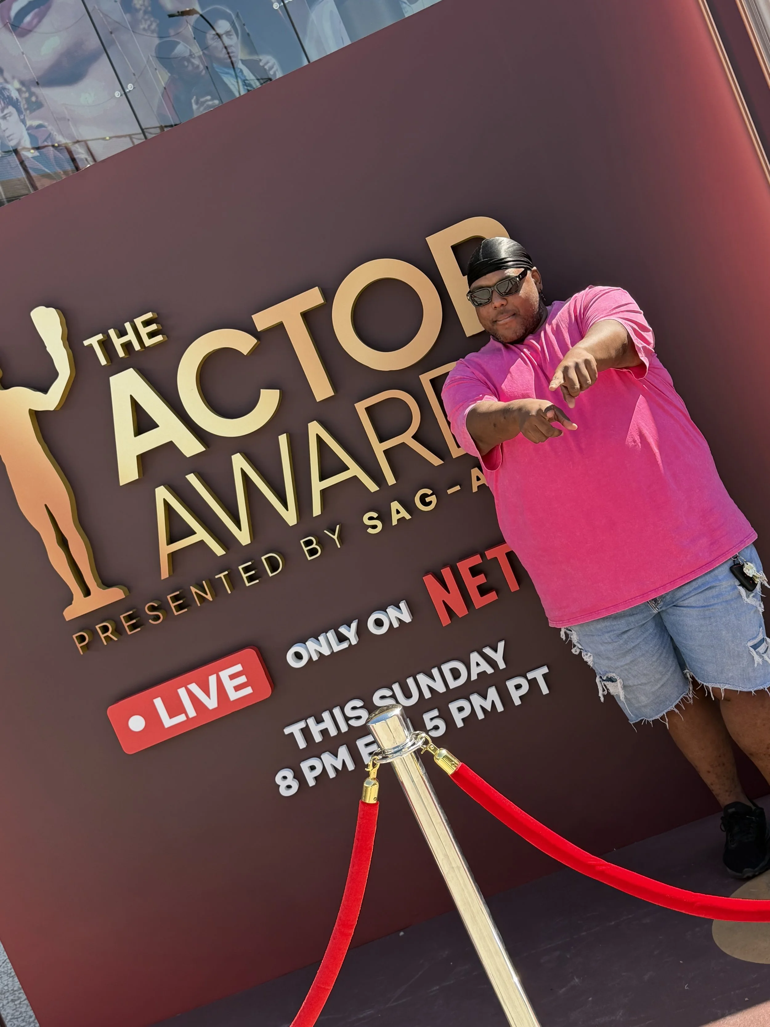 A man wearing sunglasses, a black headwrap, a pink T-shirt, and ripped denim shorts stands in front of a sign for The Acting Awards presented by SAG-AFTRA. He is pointing and aiming his hand, with a red velvet rope and stanchion in the foreground.