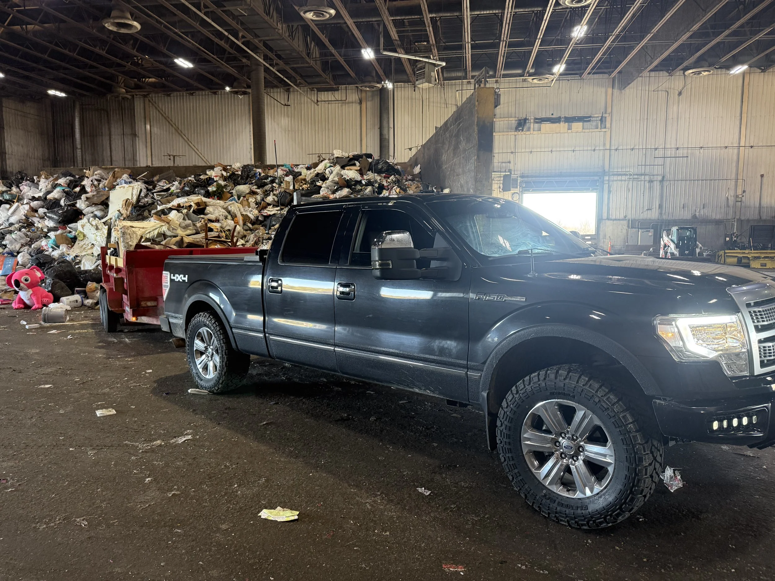 Interior of a large warehouse filled with a large pile of trash, a black pickup truck parked in front, and scattered debris on the ground.