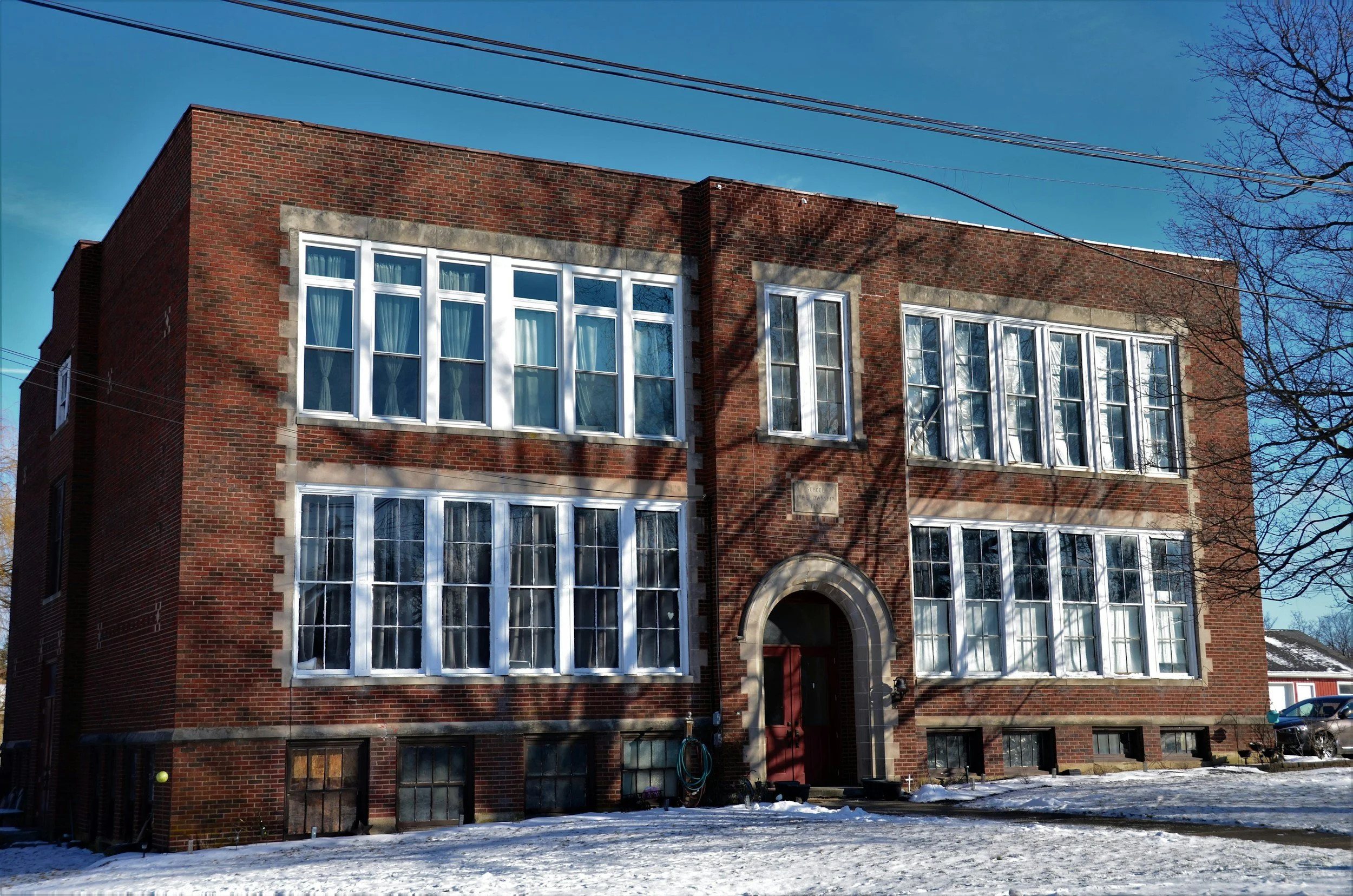 The backside of a brick school building with large white-framed windows and a red door, surrounded by snow on the ground, with a tree and power lines visible against a clear blue sky.