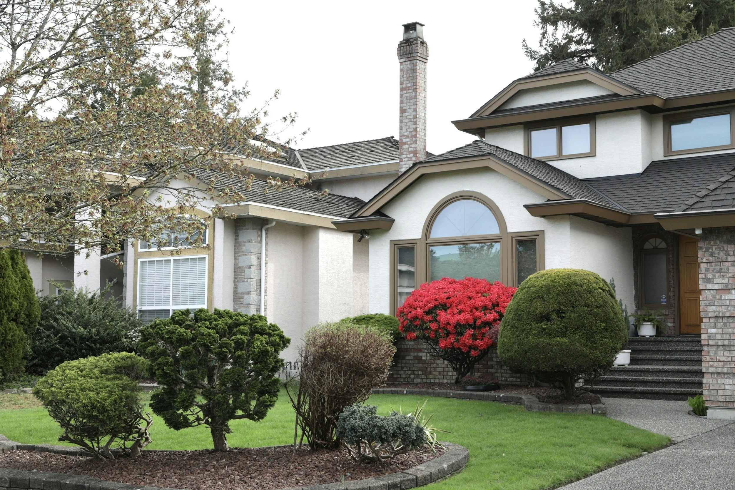 Front yard with well-manicured grass, various trimmed bushes, flowering red bush, and a sidewalk leading to a house with beige walls, large window, and a brick chimney.