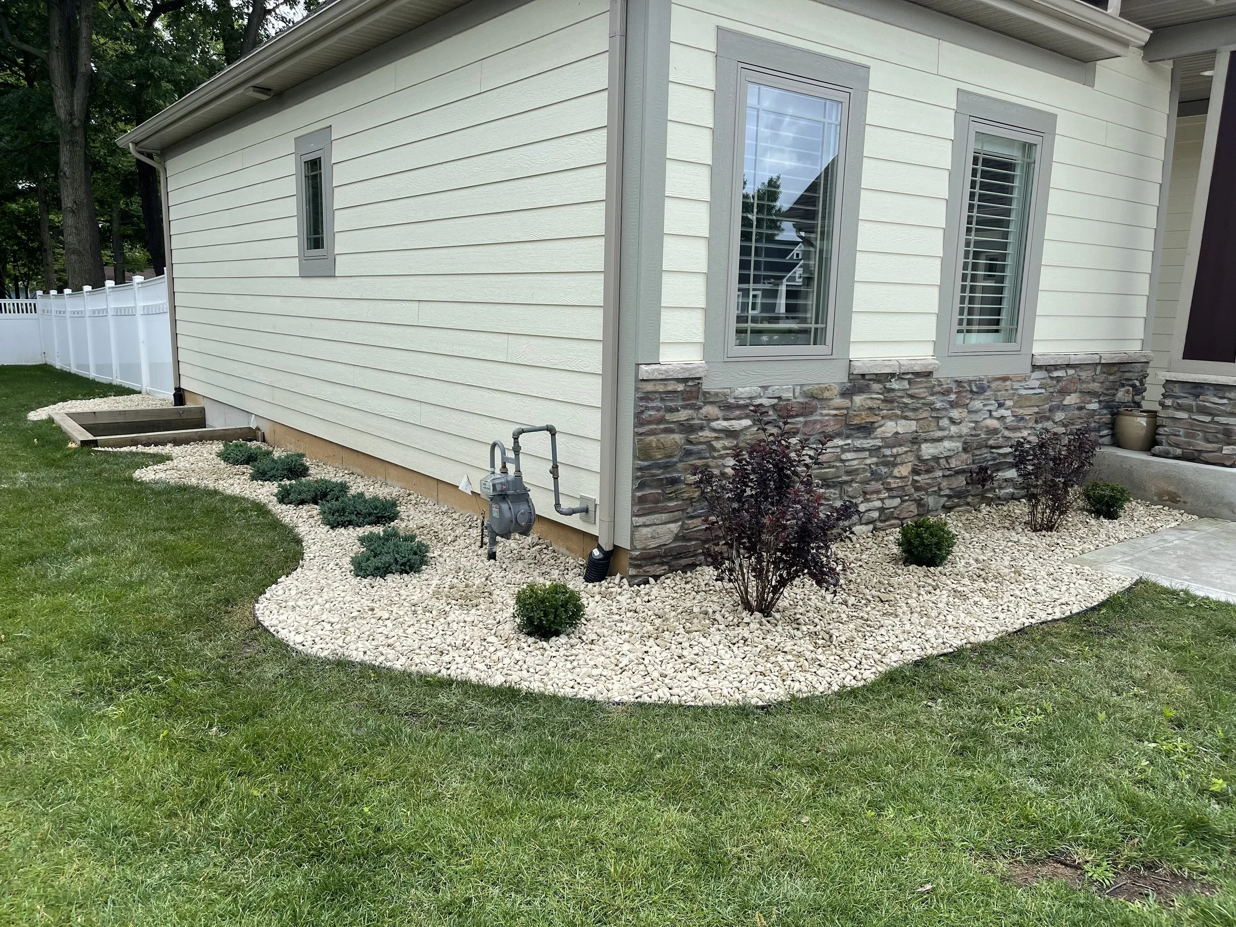 The image shows the exterior side of a house with yellow siding, three tall windows with white blinds, a stone foundation, and a small garden bed with bushes surrounded by white rocks. There is a white fence and trees in the background.