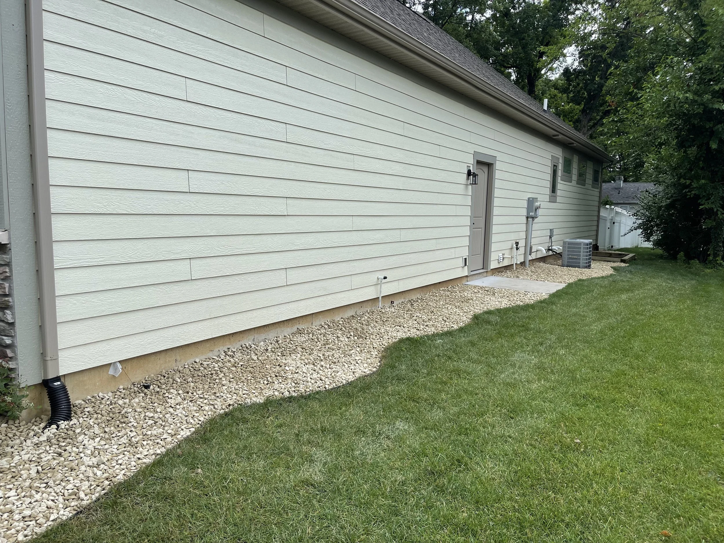 A side view of the exterior of a house with white horizontal siding. There is a small door, outdoor light, and utility connections, with gravel along the foundation and a grassy yard in Frankford, Delaware.