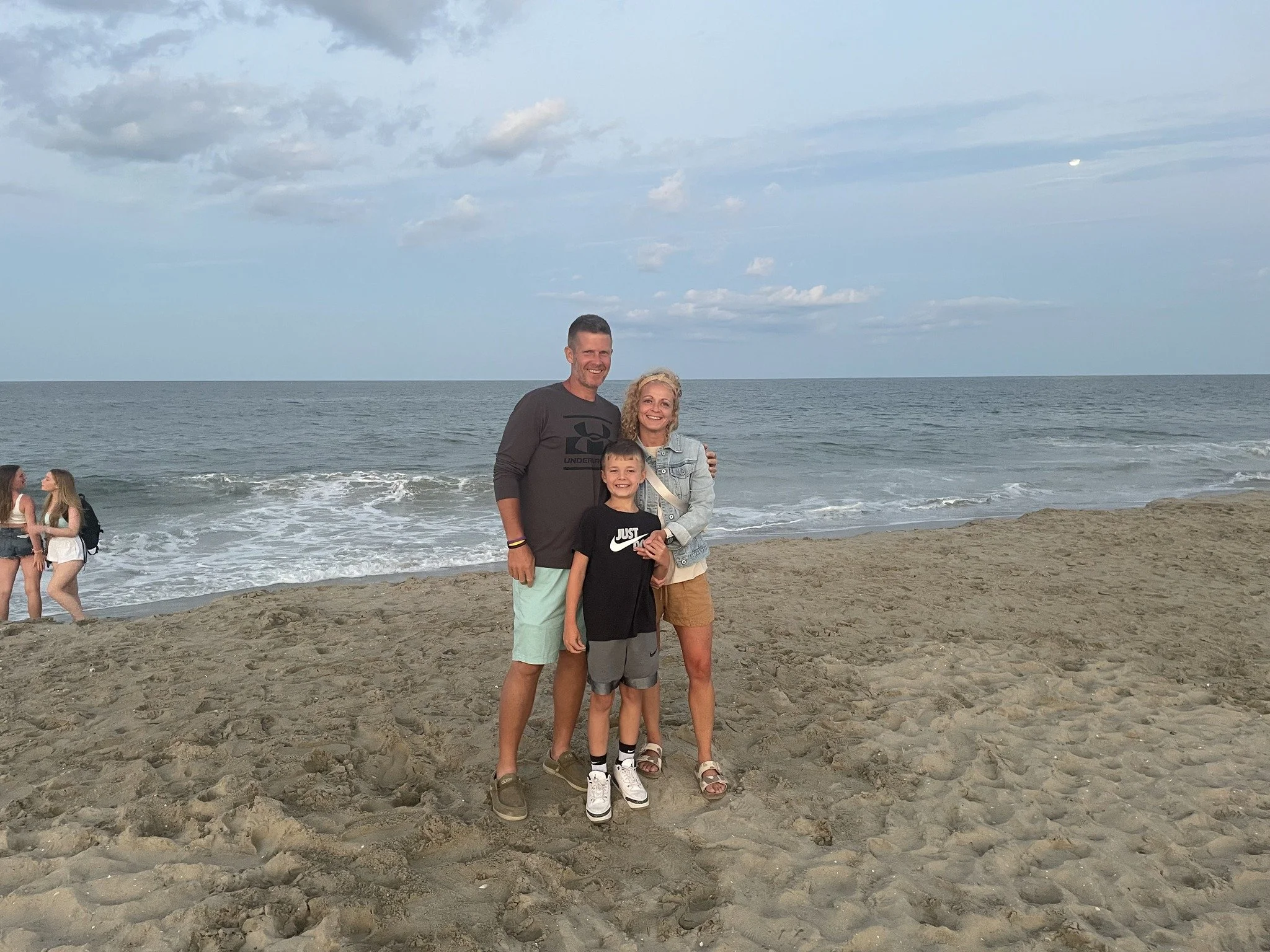 A family of three, a man, woman, and child, standing on a sandy beach with the ocean and cloudy sky in the background.