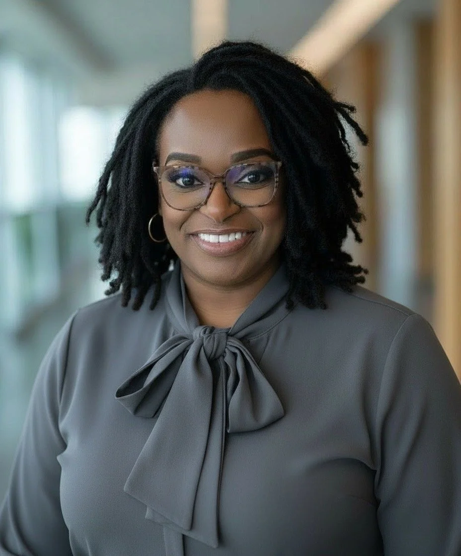 A woman with dark, curly hair, wearing glasses and a gray blouse with a bow, smiling in a well-lit indoor setting.