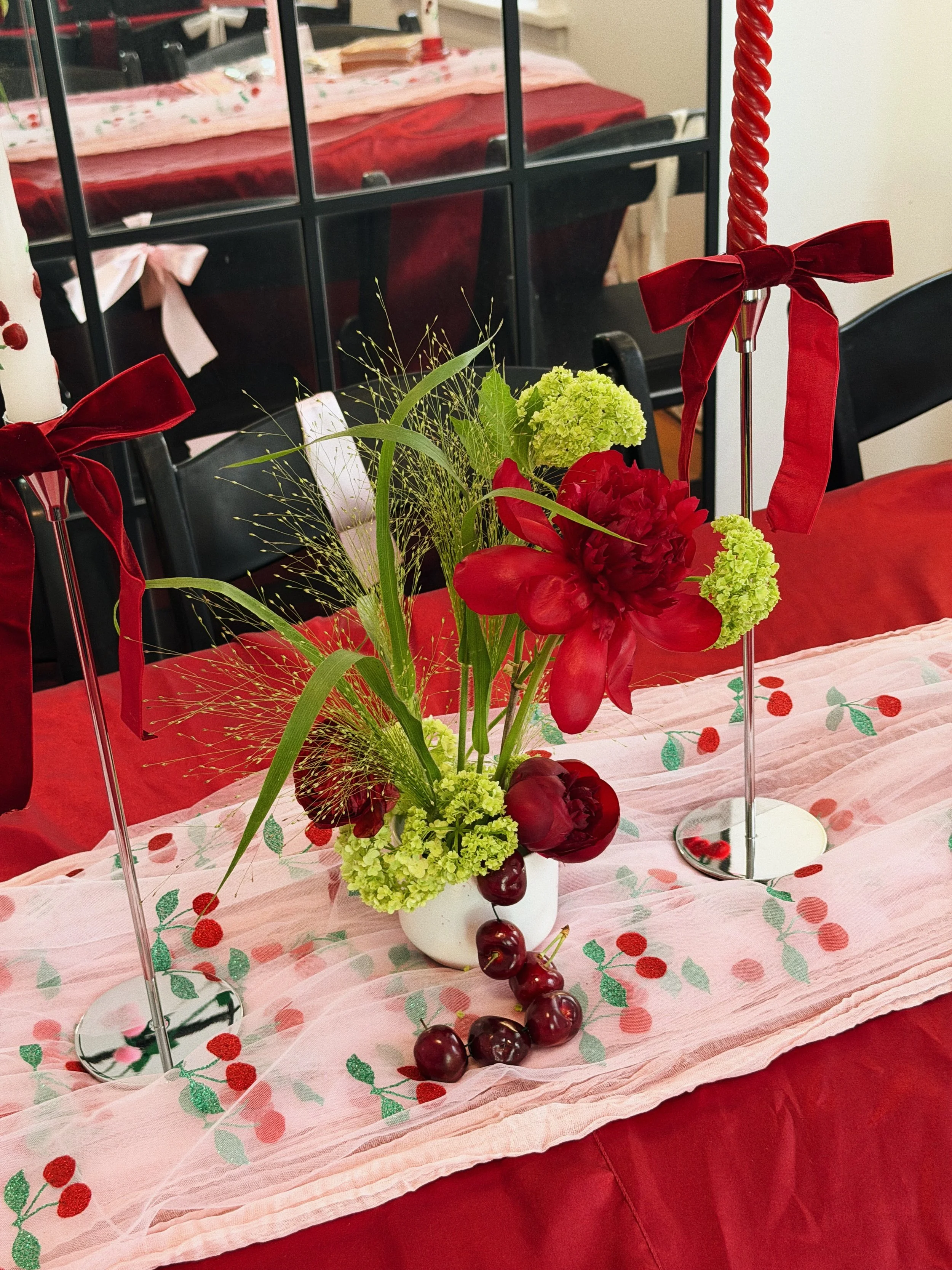 A floral centerpiece with red and green flowers on a table decorated with a cherry-patterned pink tablecloth, flanked by two tall red candle holders with red bows in a festive setting.