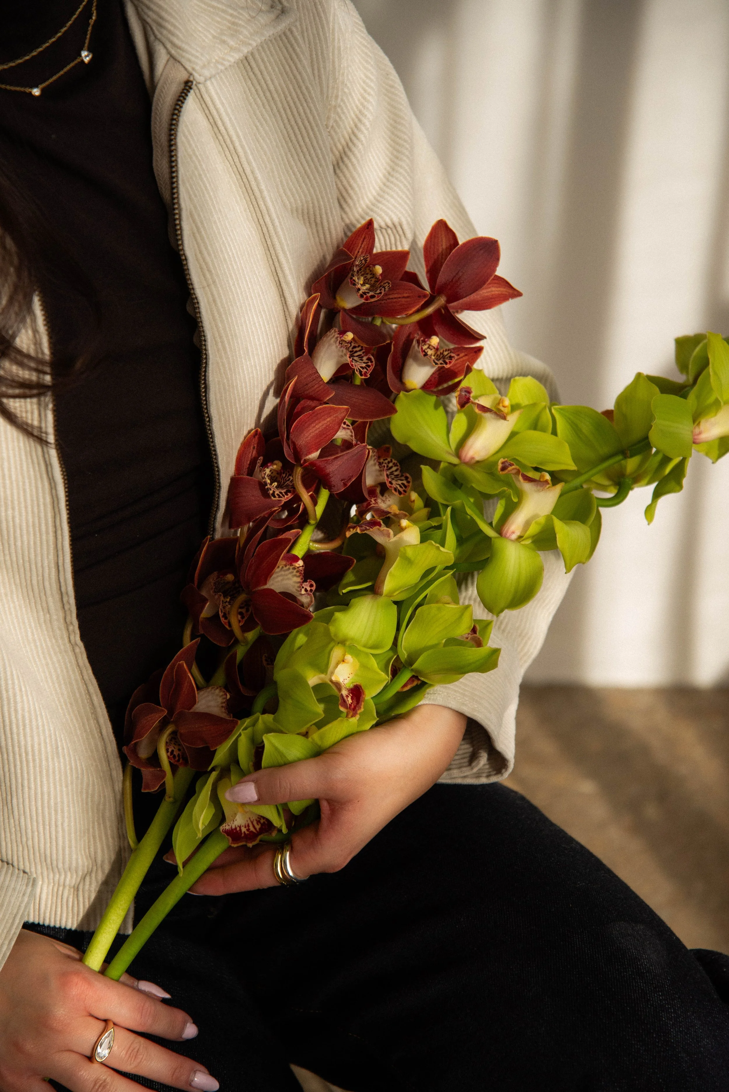 Person holding a bouquet of red and green orchids, wearing rings on fingers, black top, and a beige jacket.