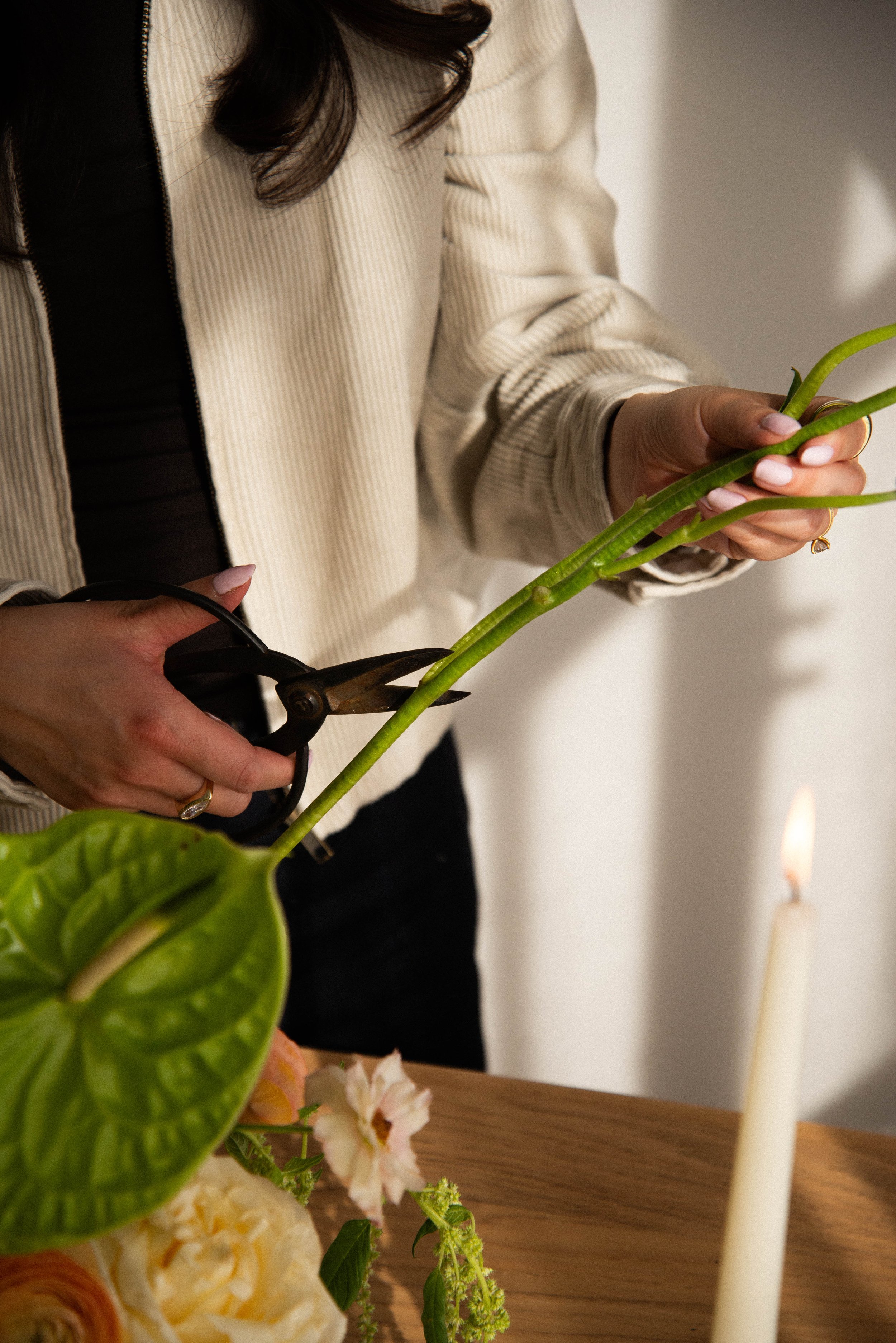 A person with dark hair wearing a beige jacket and black shirt is trimming green plant leaves with scissors, with a white candle, yellow and white flowers, and a wooden surface in the background.