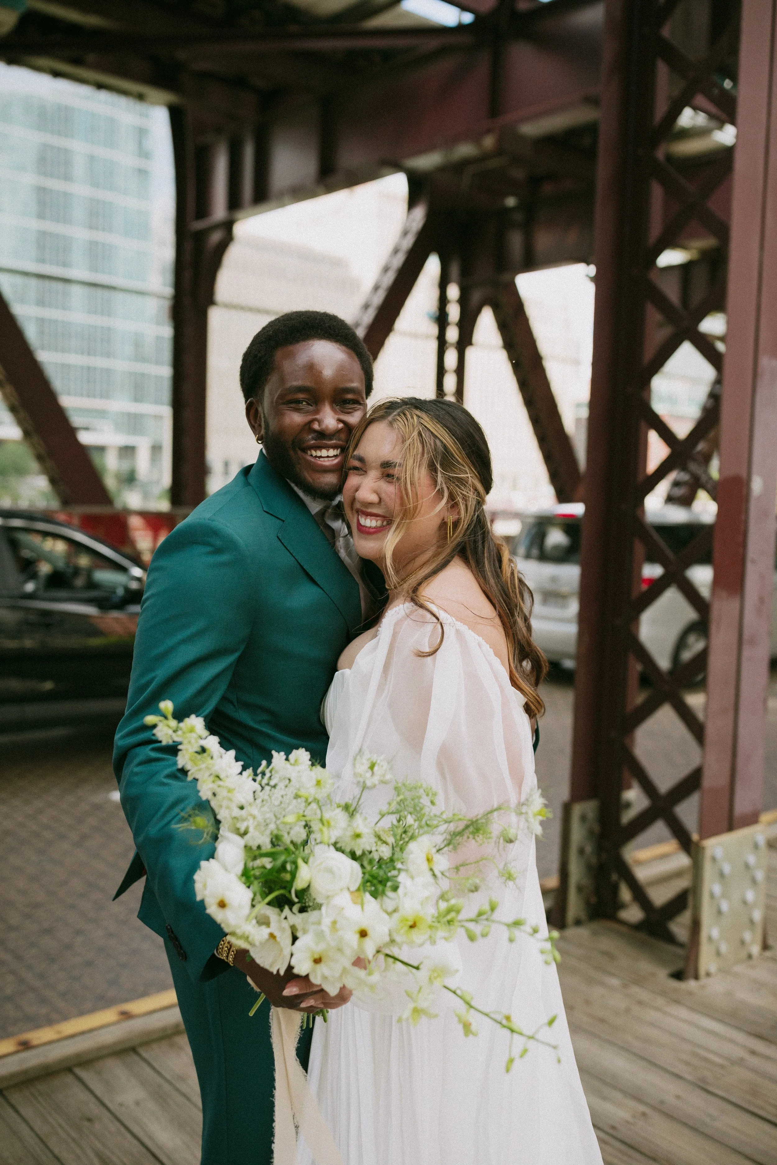 Joyful couple at a wedding, embracing and smiling, with the groom holding a bouquet of white flowers, standing underneath a metal structure near a city skyline.