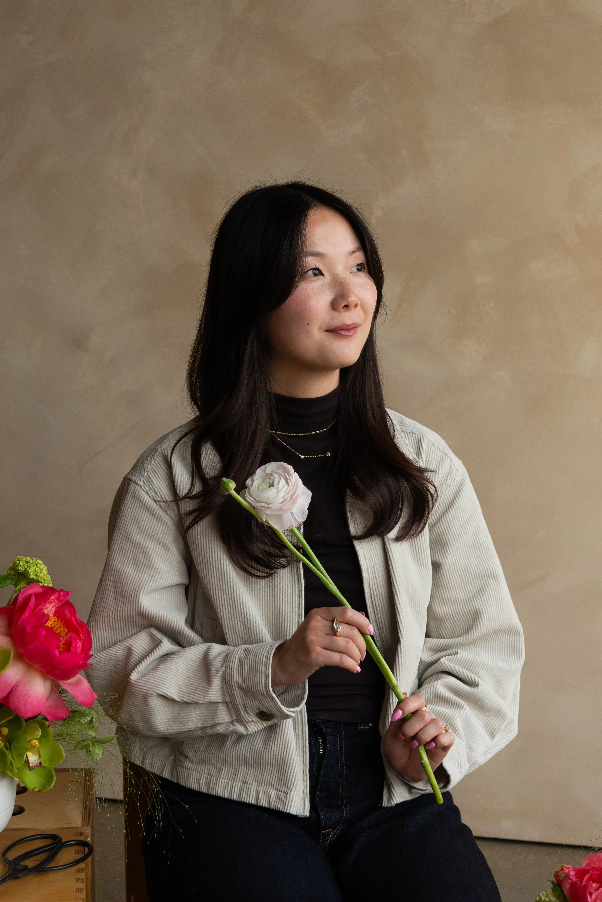 A woman with long dark hair, wearing a black shirt and a light-colored jacket, holding a white flower. She is sitting in front of a textured beige background, with colorful flowers on her left side.