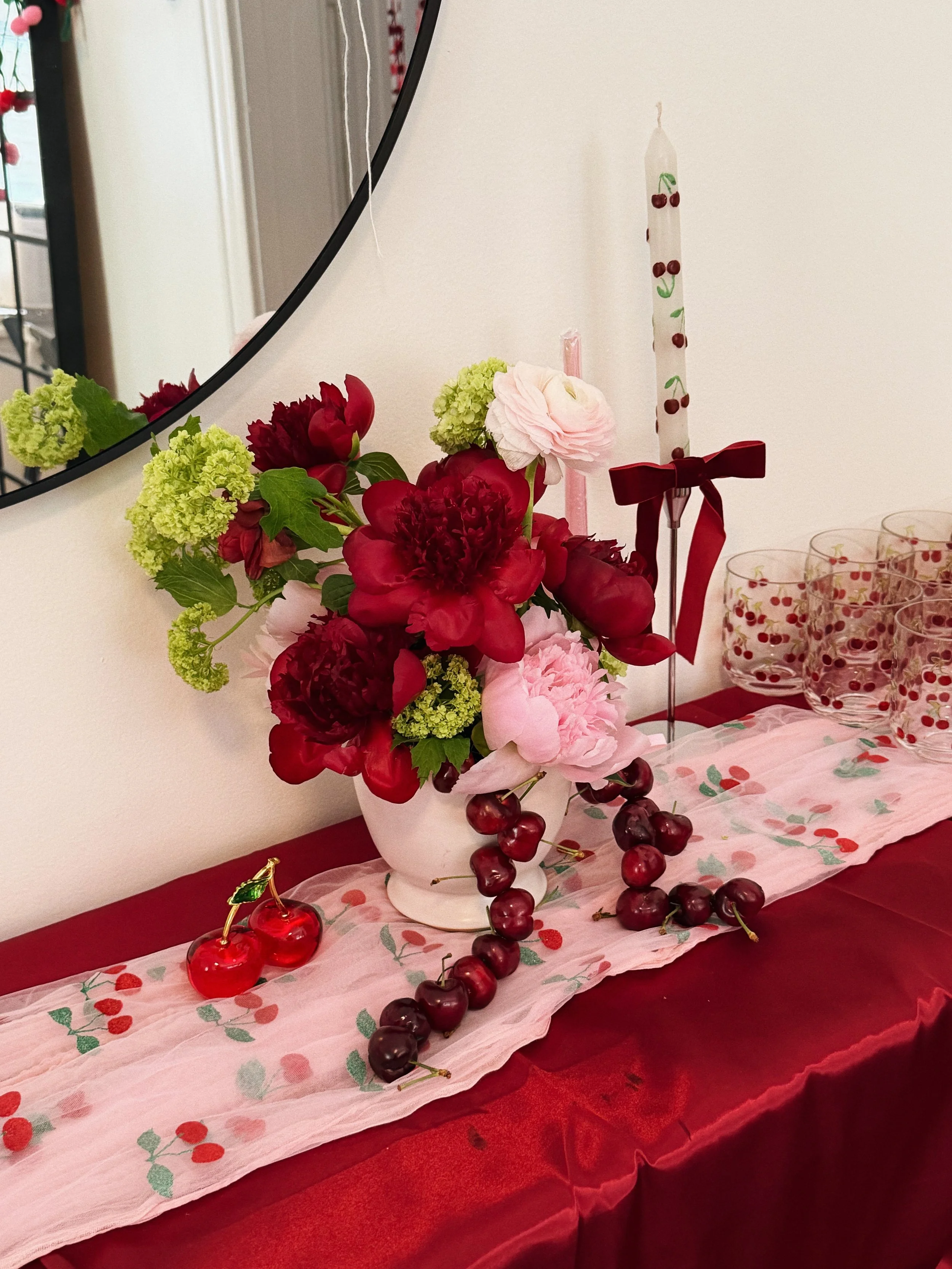 A floral arrangement with red and pink flowers in a white vase, with cherries and a cherry-themed tablecloth on a red table, alongside a tall cherry-decorated candle and glassware.