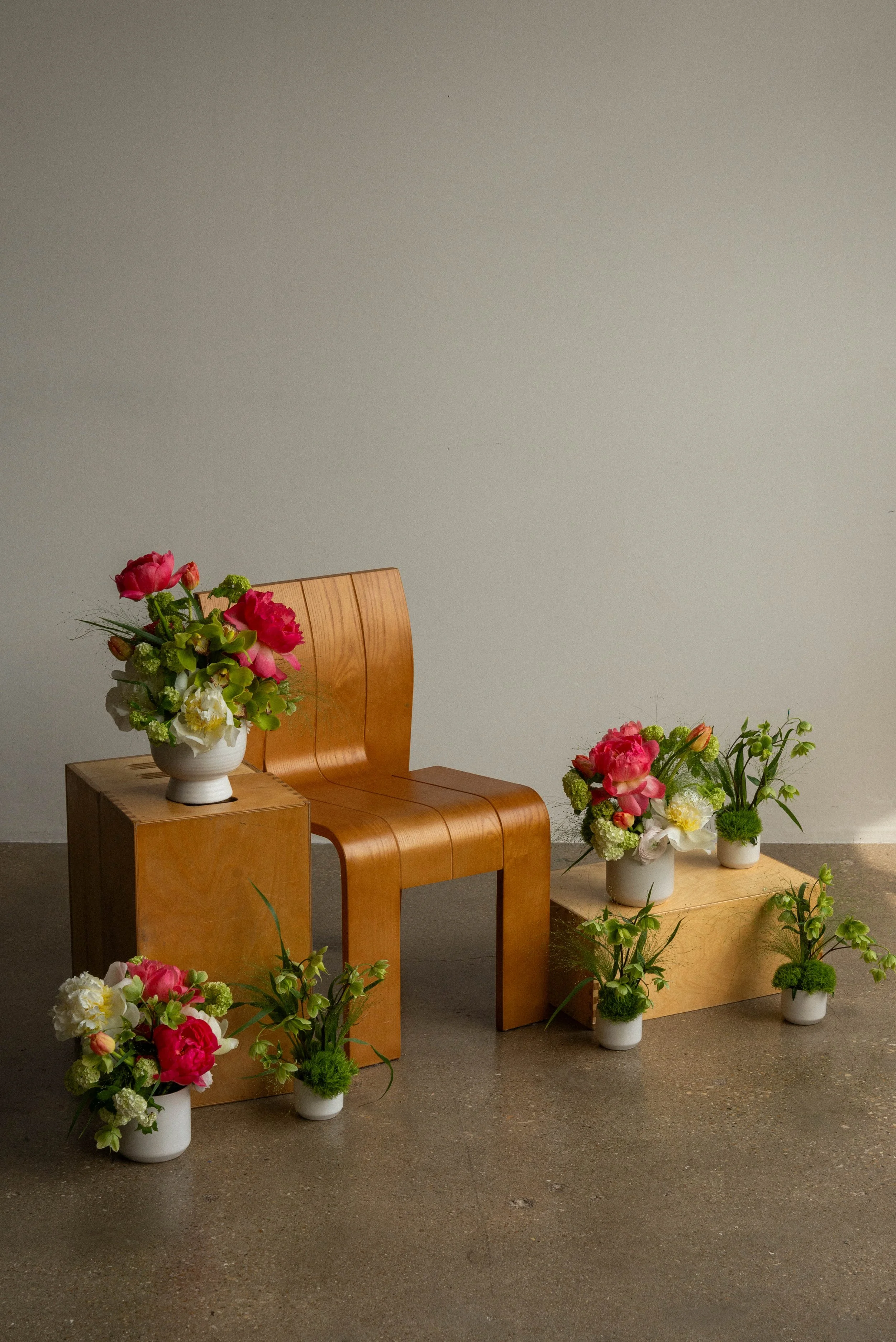 Several flower arrangements with pink, white, and green flowers in white vases, placed on wooden furniture against a plain gray wall.