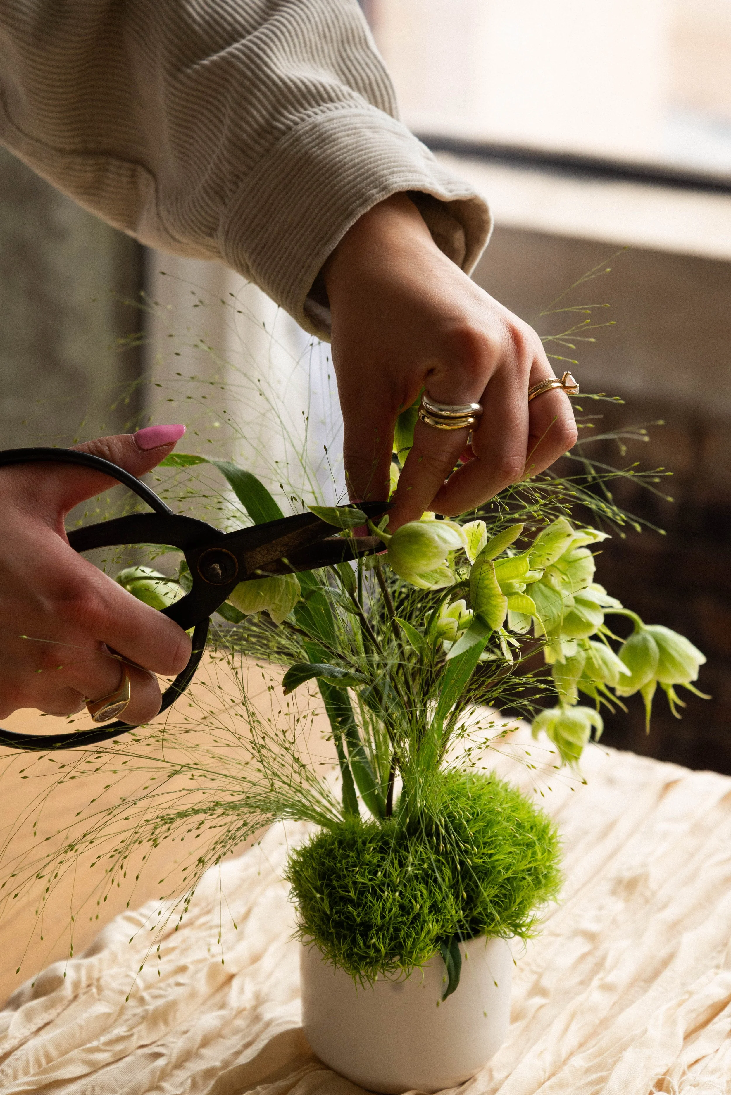 A person arranging a flower arrangement with scissors in a white vase, featuring green and yellow flowers and moss, on a cream-colored fabric surface.
