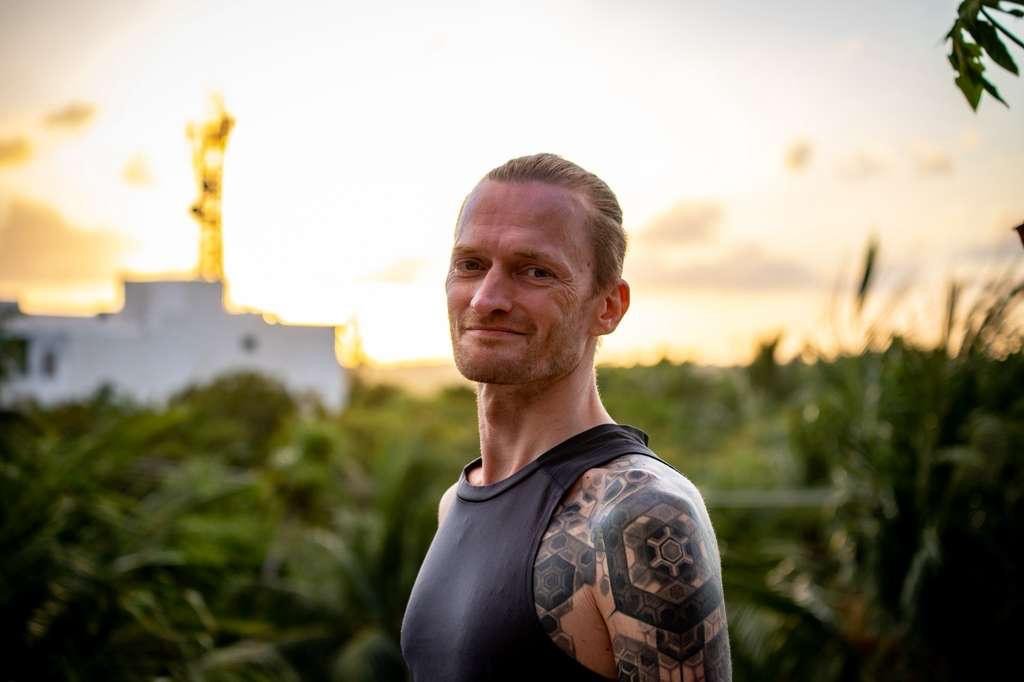 A man with tattoos on his left arm standing outdoors at sunset, smiling, with greenery and a white building in the background.