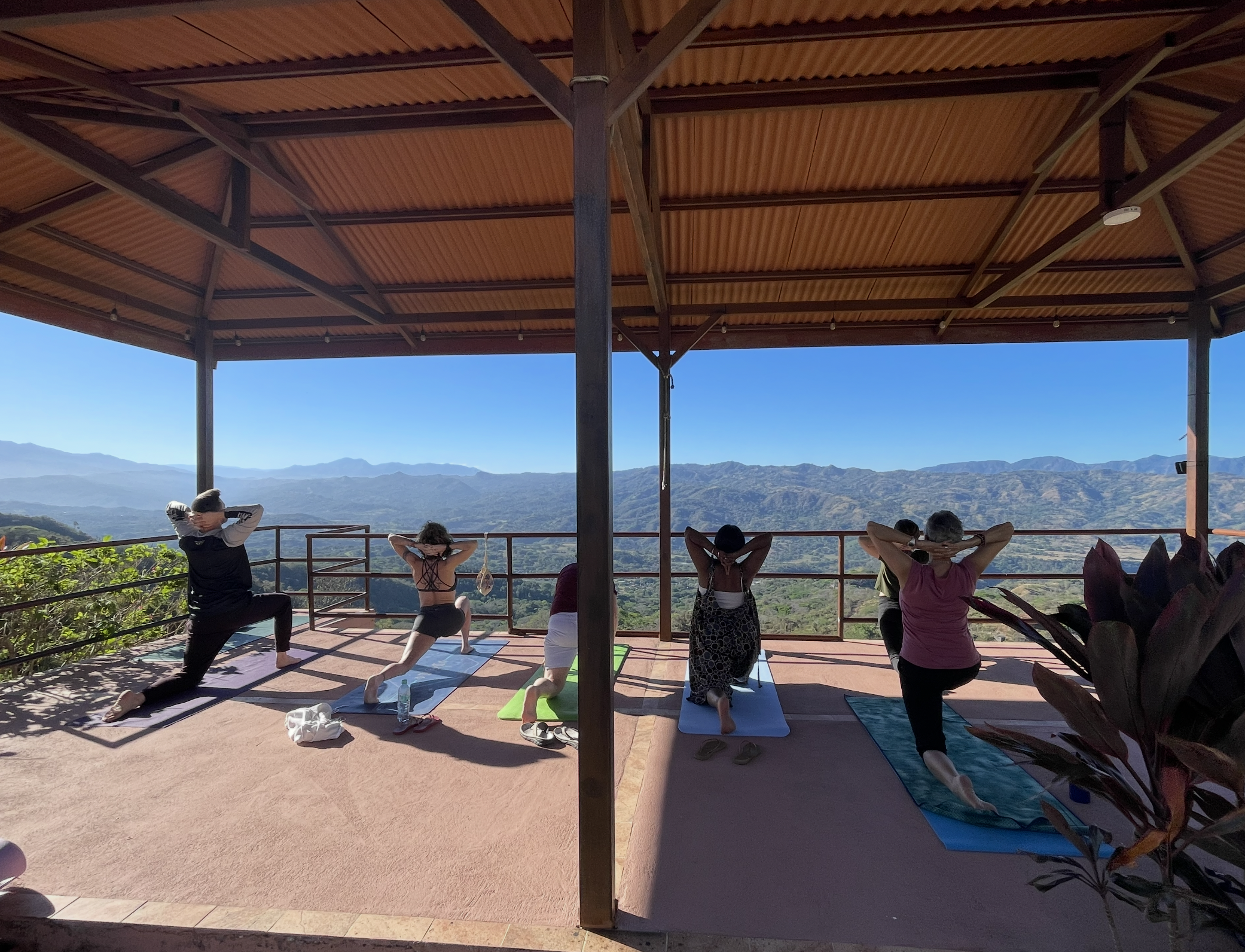 People practicing yoga outdoors on mats under a large open pavilion, with mountain scenery in the background.