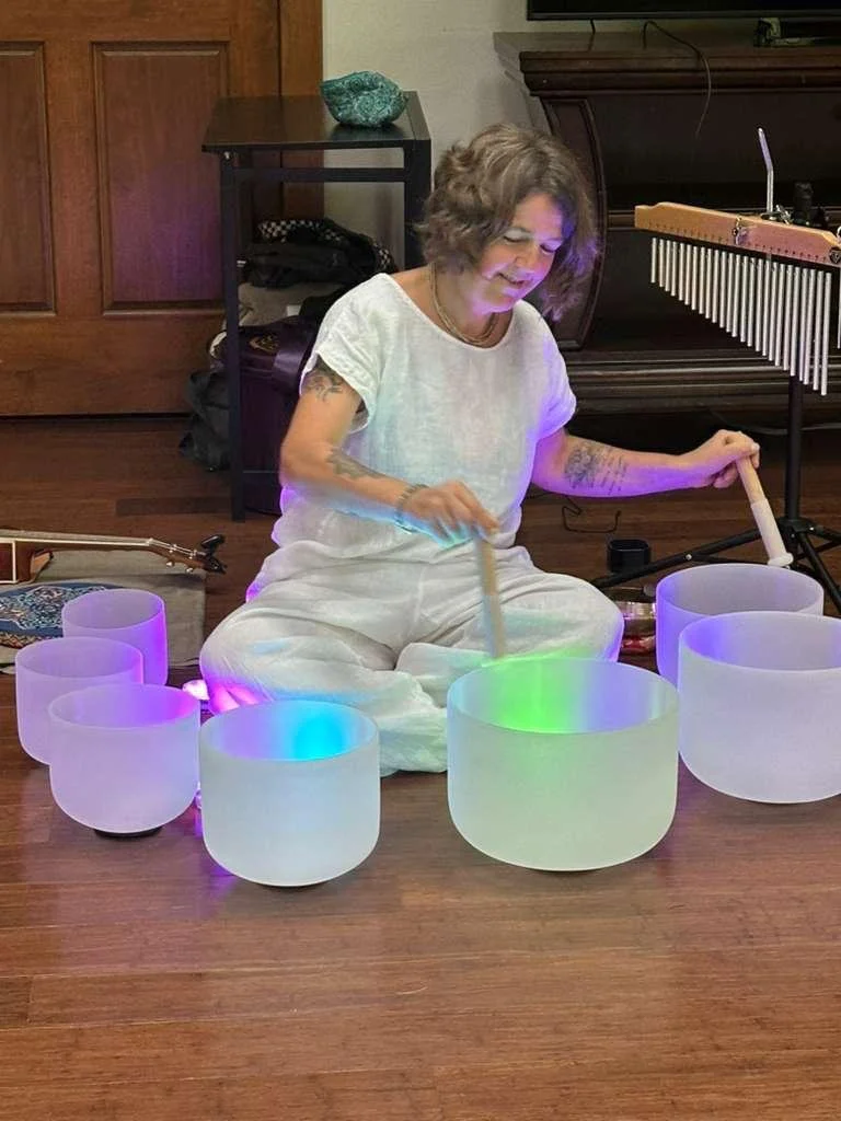A woman sitting on the floor playing a set of illuminated crystal singing bowls with a mallet in a room with wooden floors and furniture.