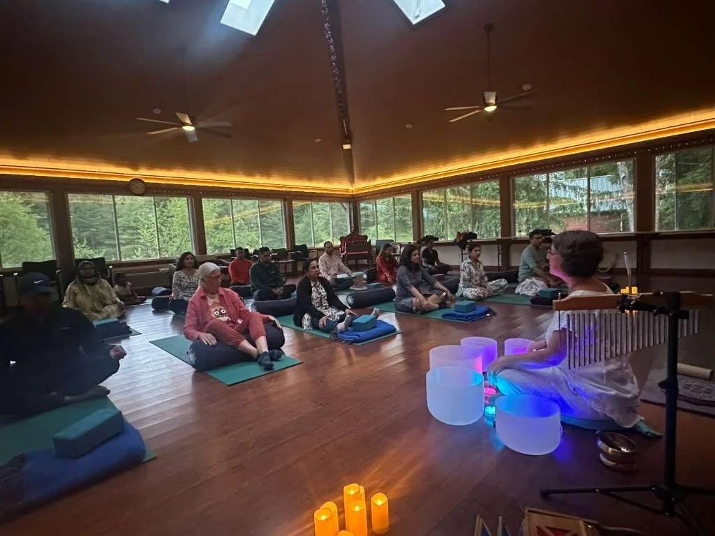 A group of people participating in a yoga or meditation class in a spacious room with large windows, wood floor, and ambient lighting. The instructor is seated with lit candles and colorful illuminated bowls in front.