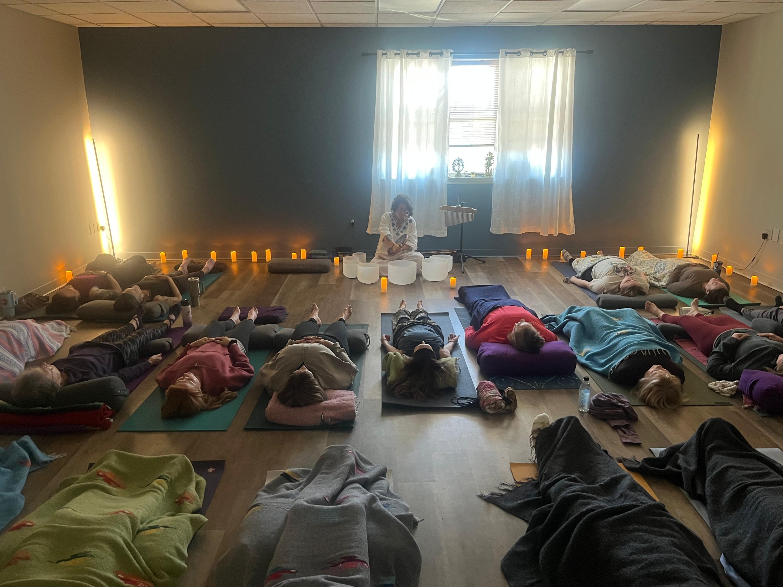 A group of people lying on their backs on mats in a room, participating in a group meditation or relaxation session, with a woman sitting cross-legged playing singing bowls at the front near a window with sheer curtains, decorated with small statues,
