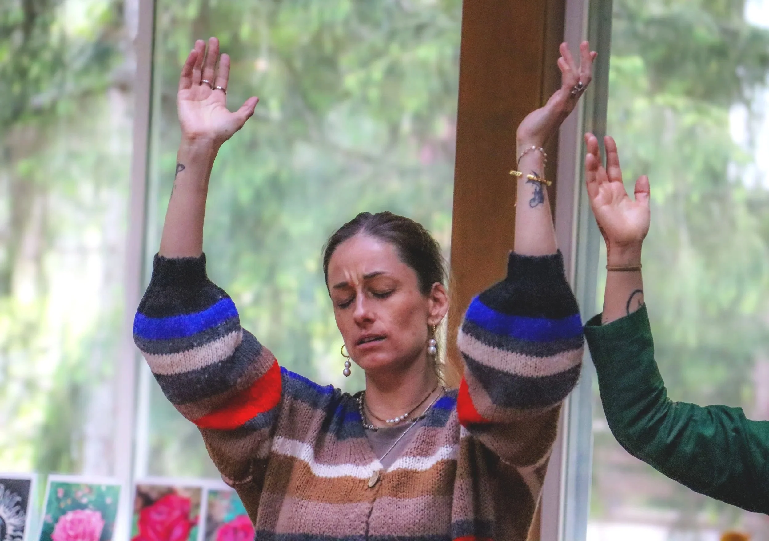 A woman with three tattoos on her arms, wearing a striped sweater and jewelry, has her eyes closed and appears to be in a moment of emotion or concentration. She is raising her hands, and other people are partially visible around her. The background 