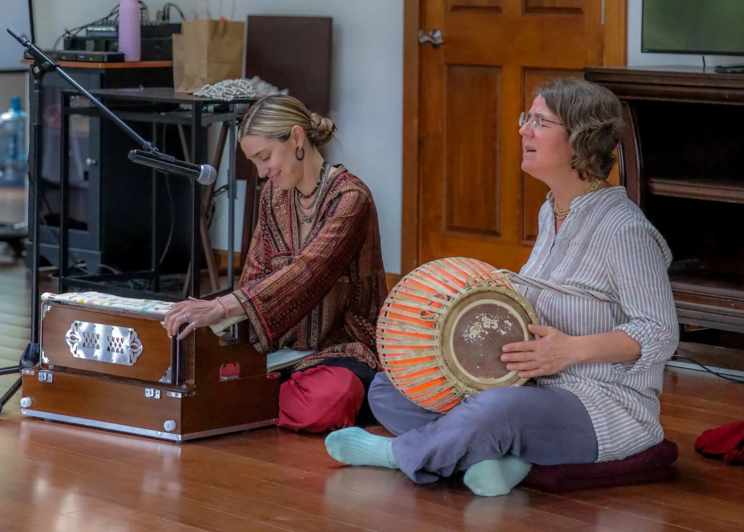 Two women are sitting on the floor during a musical performance. One woman is playing a small wooden instrument, and the other woman is holding a traditional drum. Both women are smiling and engaging with each other in a room with wooden floors and a