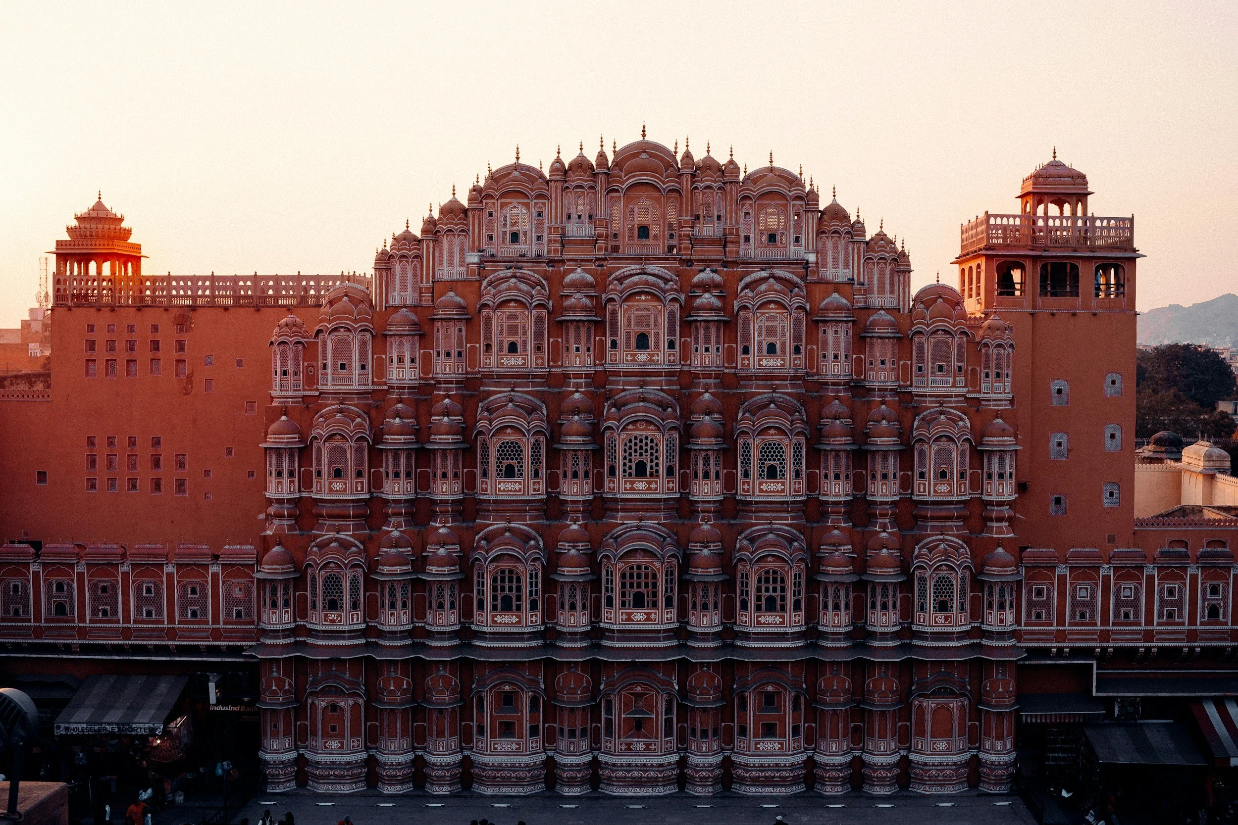 View of a grand, ornate red sandstone building with intricate white detailing, multiple domes, and minarets, likely an Indian palace or historical monument, during sunset.