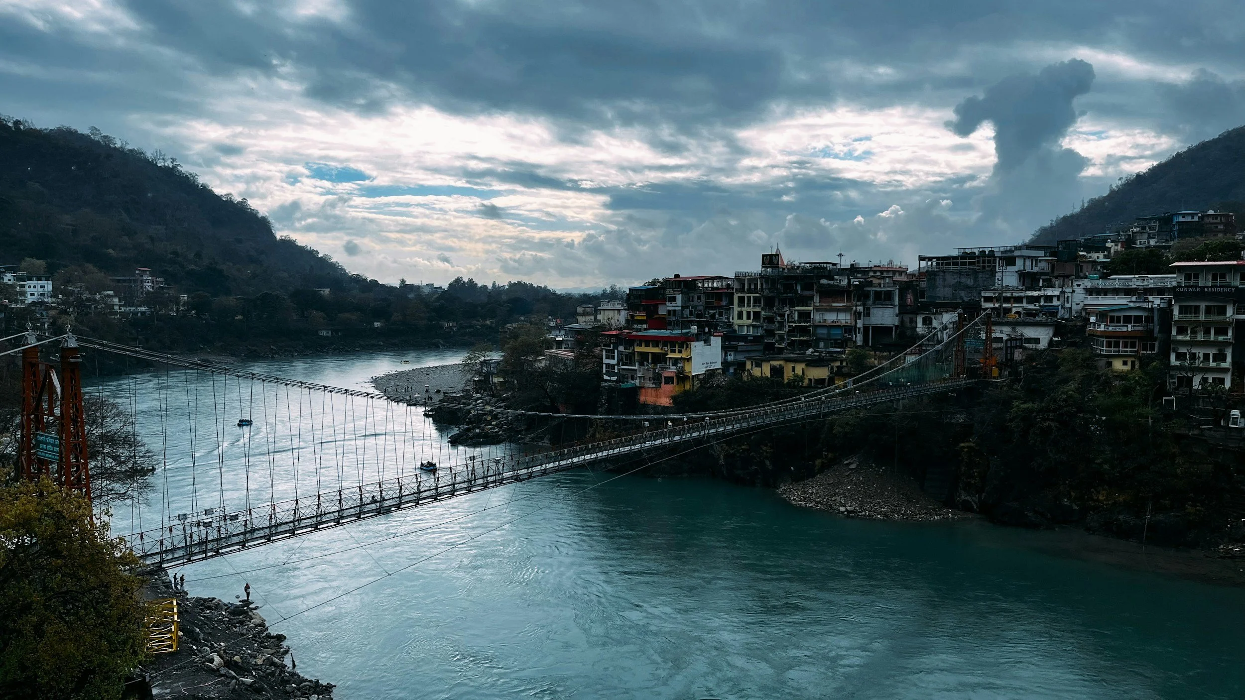 A river flowing through a valley with a suspension bridge crossing over it. On the hillside, there are buildings and houses, with cloudy skies overhead.