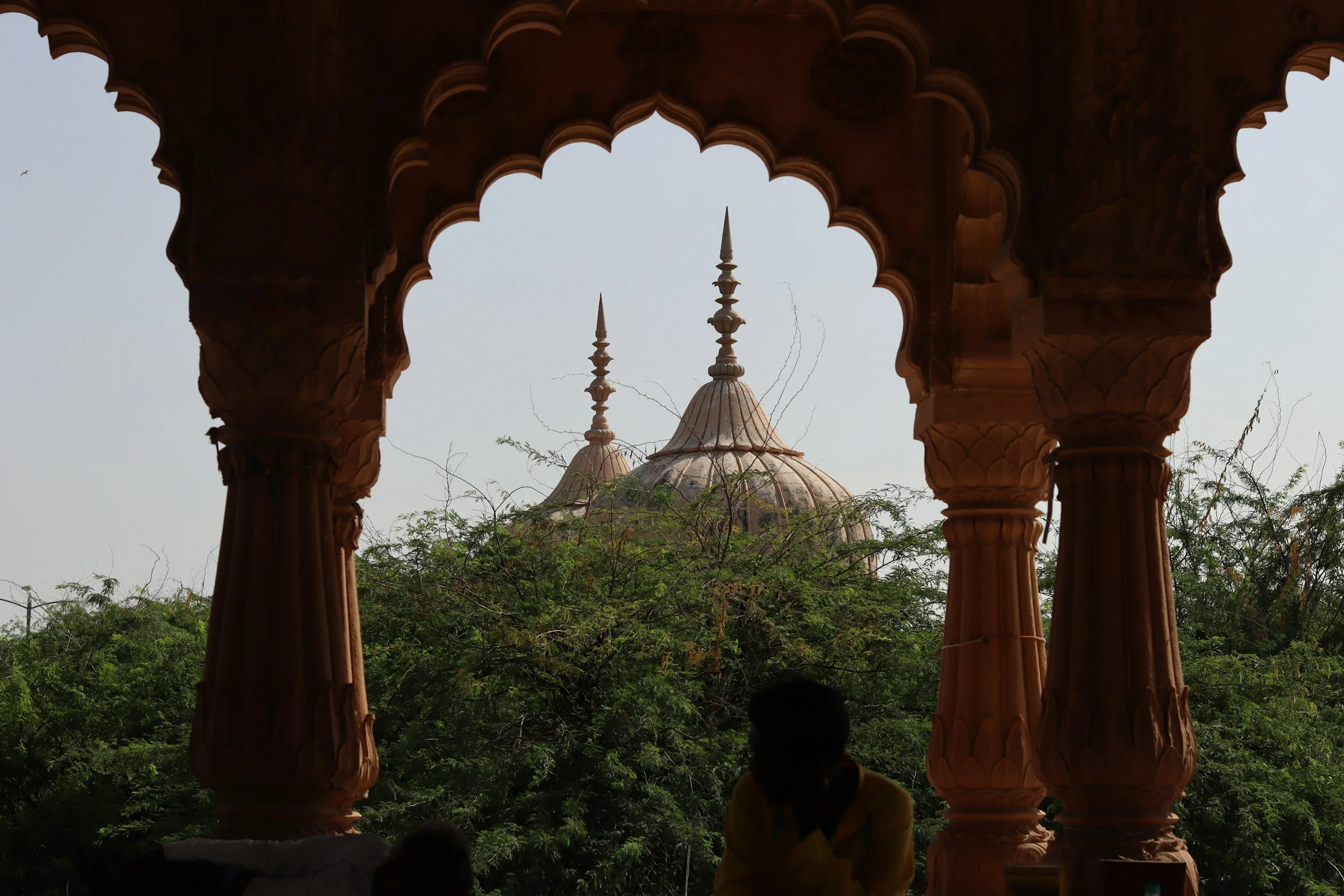View of a historic building with domes and spires through ornate arches of a balcony or pavilion, surrounded by lush green foliage.