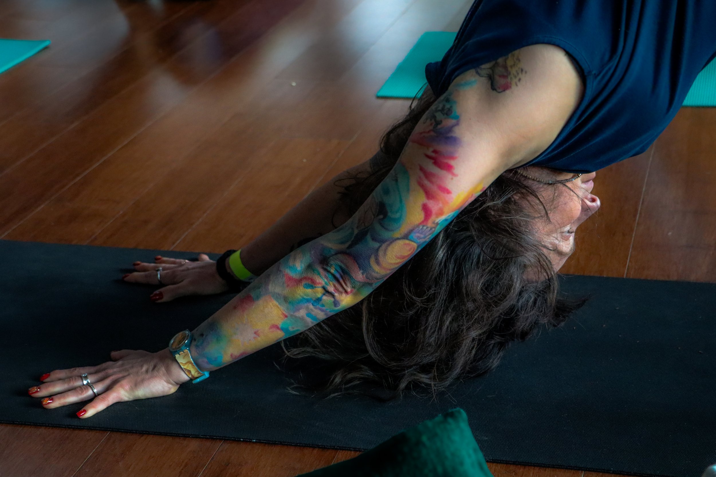 A woman with tattoos practicing yoga in downward dog pose on a black yoga mat in a wooden-floored room.